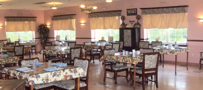 A dining room with multiple tables covered in floral tablecloths, each set with glasses and napkins. The room has pink walls, large windows with cream-colored valances, and upholstered chairs with floral patterns. There is a cabinet with decorative items and a sign above it in the background.