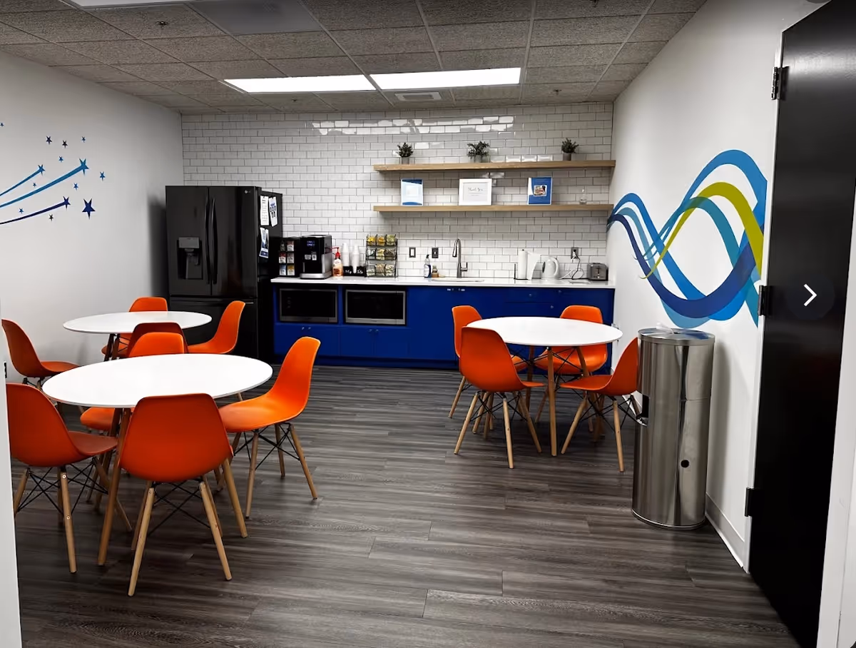 Bright communal dining area with round white tables, orange chairs, and a blue kitchenette.