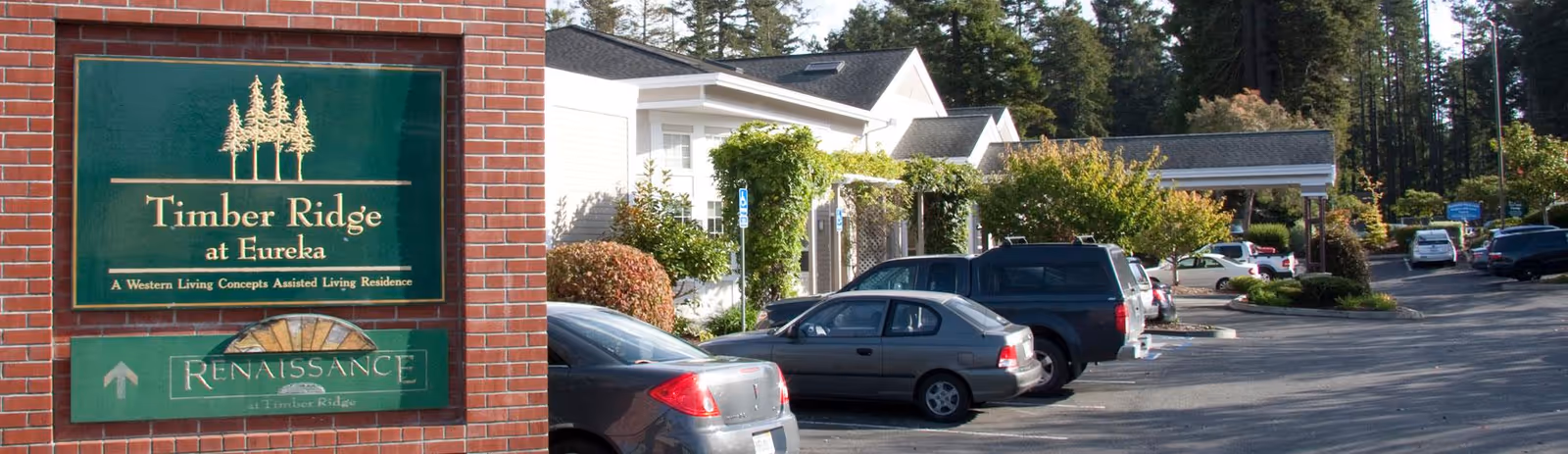 Brick entrance sign reading 'Timber Ridge at Eureka' with parked cars and the facility's front entrance in the background.