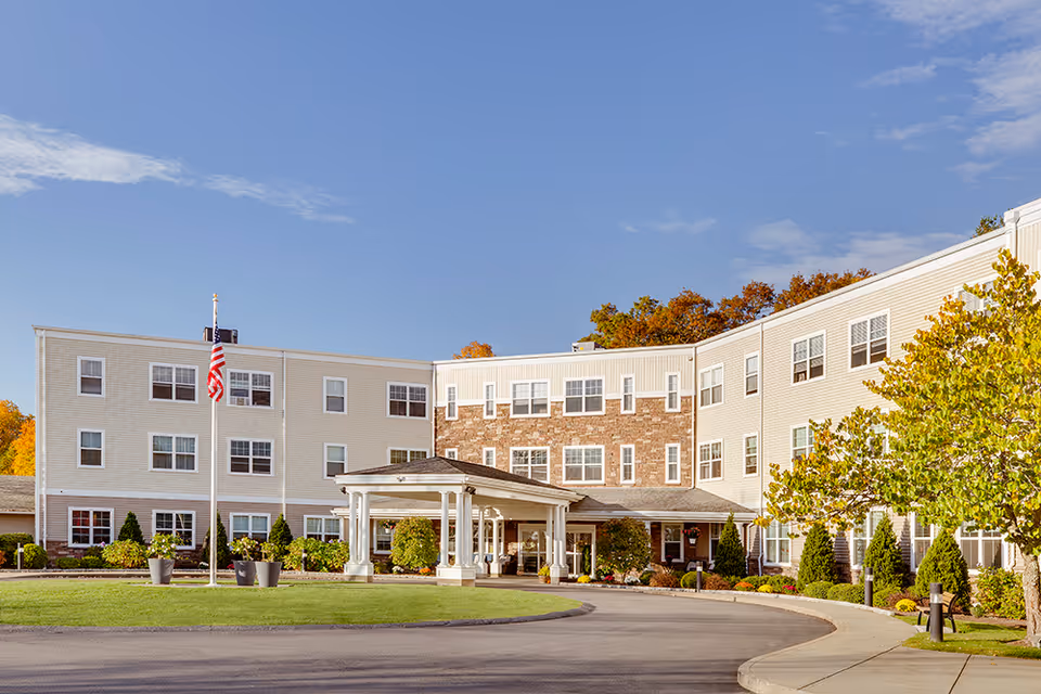 Exterior view of The Residence at Great Woods, a three-story senior living facility with beige siding and stone accents. The building has many windows and a covered entrance with white columns. There is a curved driveway, green lawn, trees, and an American flag on a flagpole in front of the building under a clear blue sky.