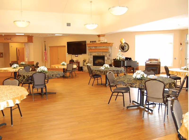 Spacious dining/activity room with round tables and chairs, a fireplace, wall-mounted TV, and a piano.