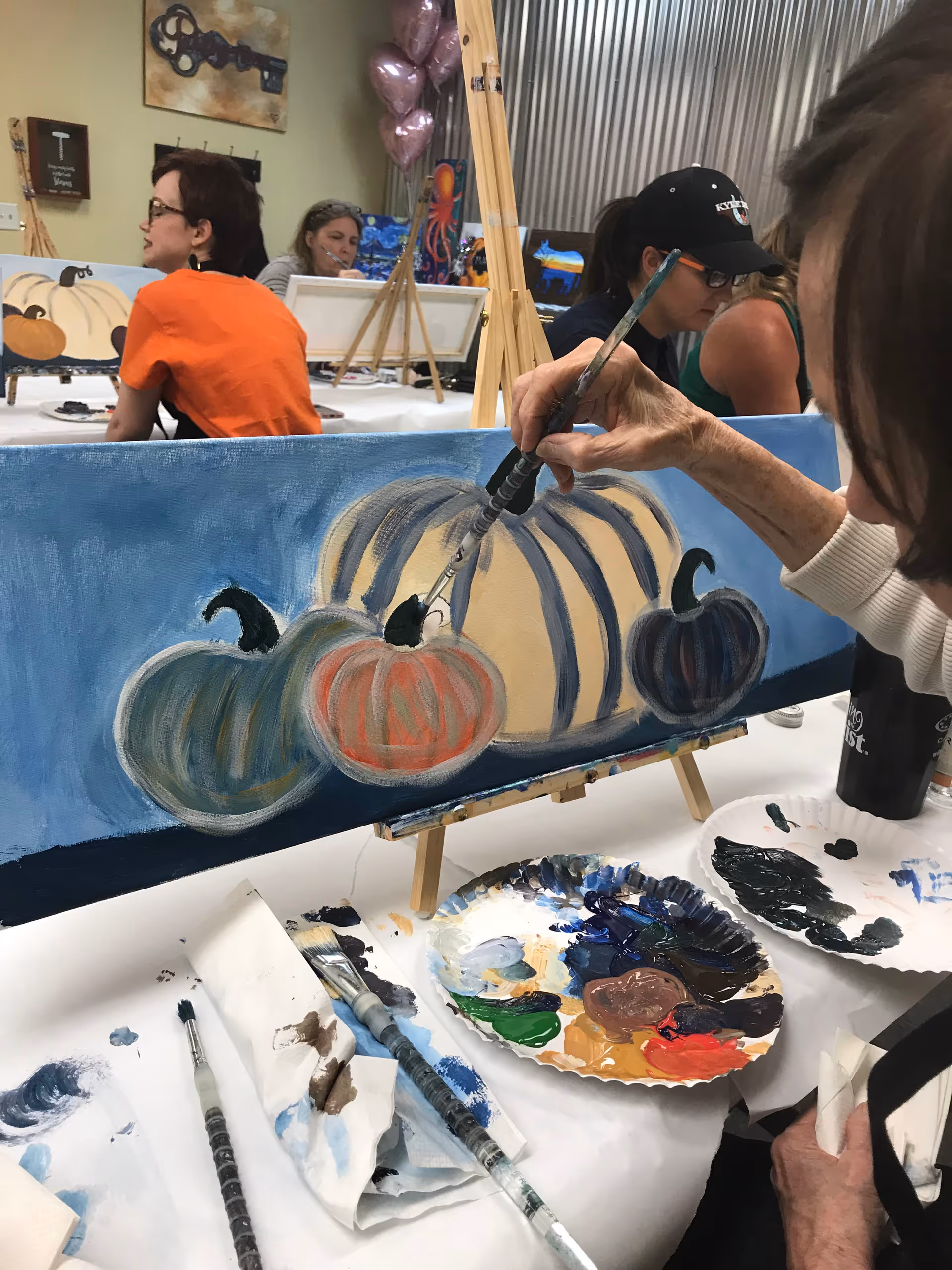 Indoor art class showing a pumpkin painting on a blue canvas with supplies nearby.