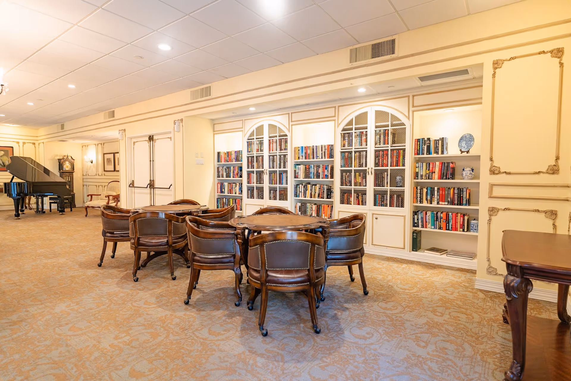 A spacious and elegant interior room with a carpeted floor, featuring a round wooden table surrounded by six leather chairs. The back wall has built-in bookshelves filled with books and decorative items. To the left, there is a grand piano and a tall grandfather clock. The room has soft lighting and classic decorative molding on the walls.