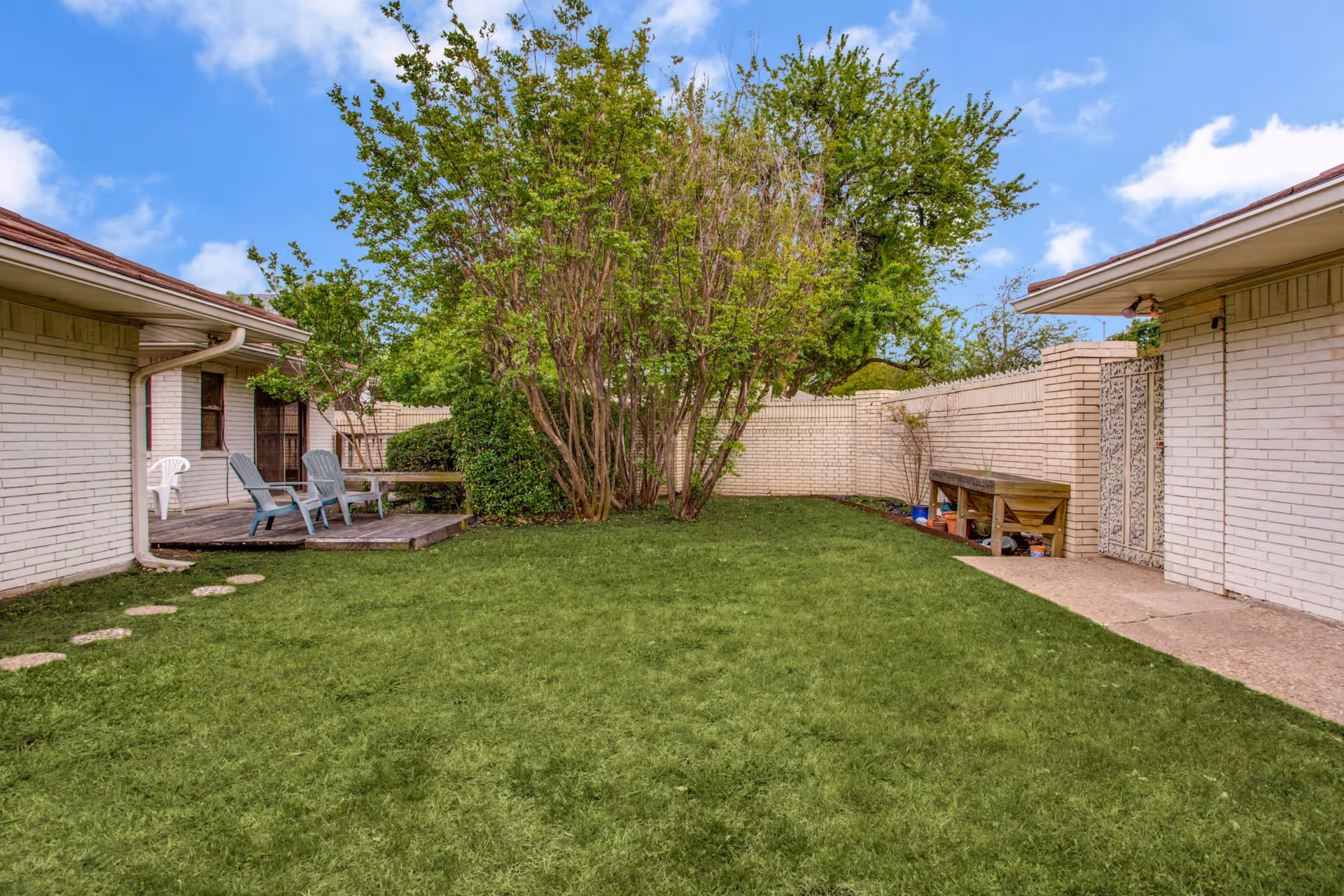 A grassy enclosed courtyard with a wooden deck and chairs, trees, and light brick building walls under a blue sky.