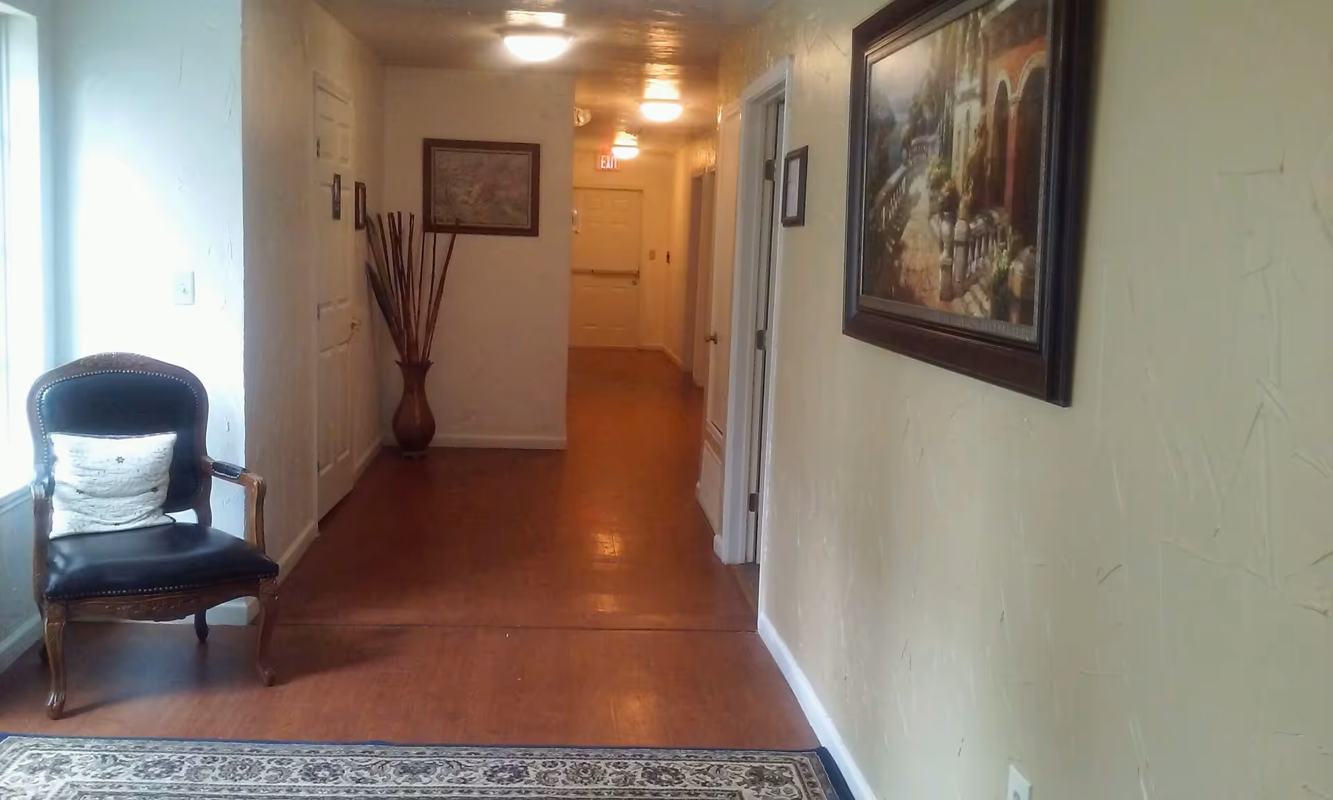 A hallway in an assisted living facility with wooden flooring and cream-colored textured walls. There is a black cushioned chair with a white pillow on the left near a window, a decorative vase with tall sticks at the end of the hallway, and framed paintings on the walls. The hallway is lit by ceiling lights and has several doors along the right side.