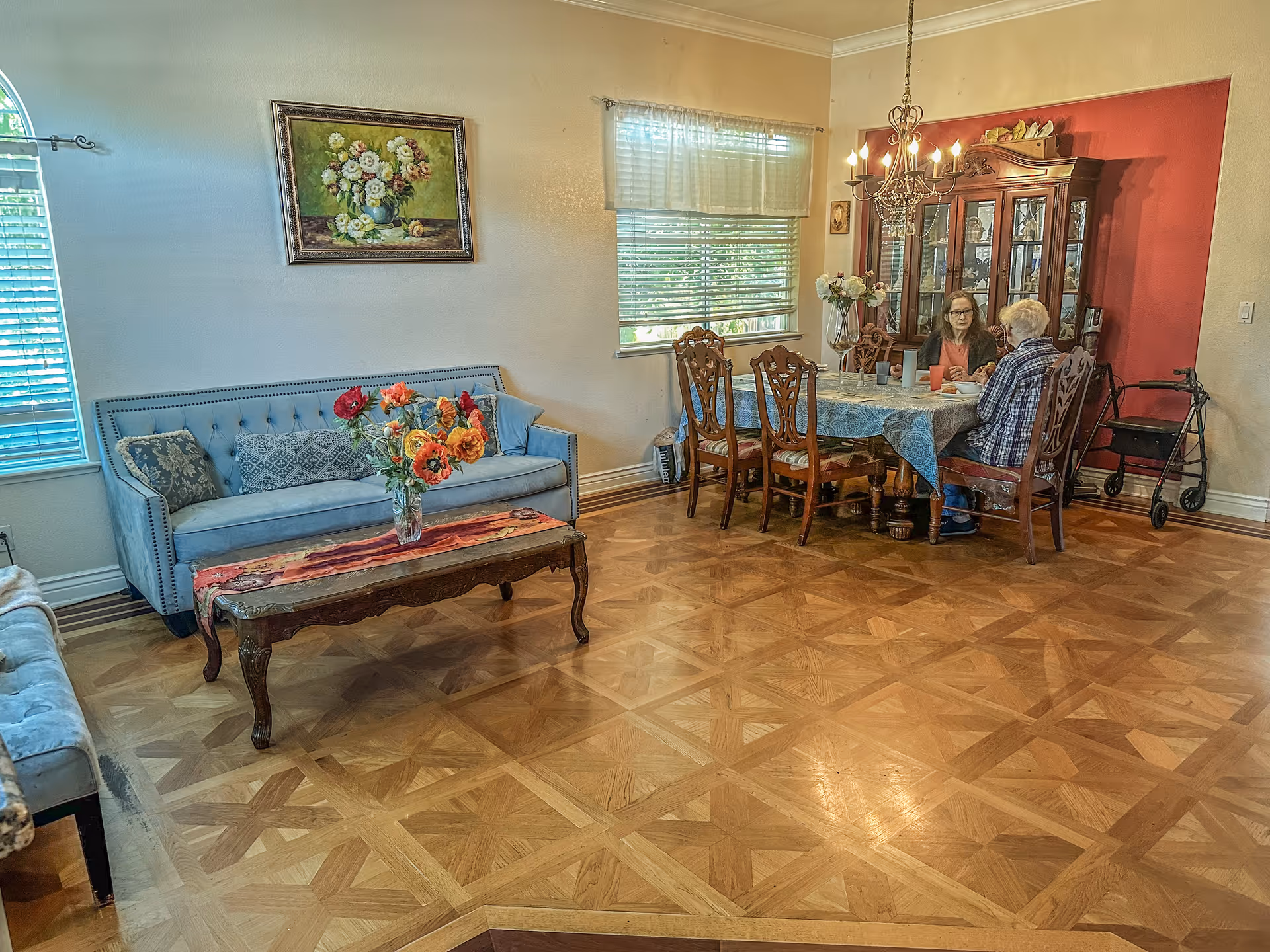 A cozy living-dining room with a blue sofa, flowers, and people at a dining table.