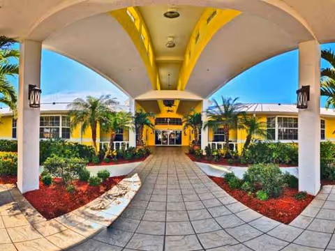 Wide covered entrance leads to a yellow building with palm trees and a walkway.