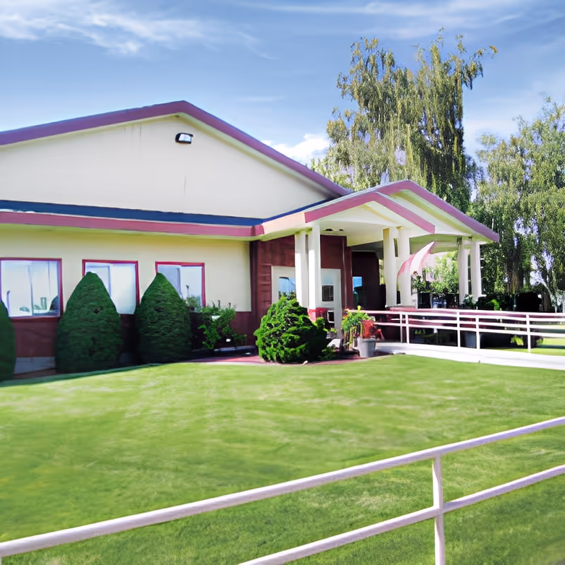 Front exterior of a single-story senior living facility with a covered entrance, wheelchair ramp, manicured lawn, and shrubs.