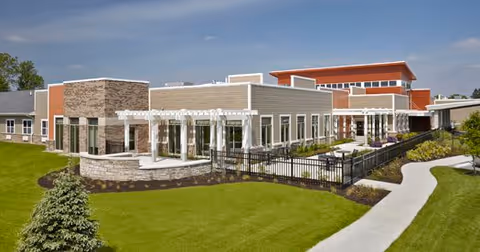 Exterior view of a modern senior living facility building with a well-maintained lawn, paved walkway, and a fenced patio area under a clear blue sky.
