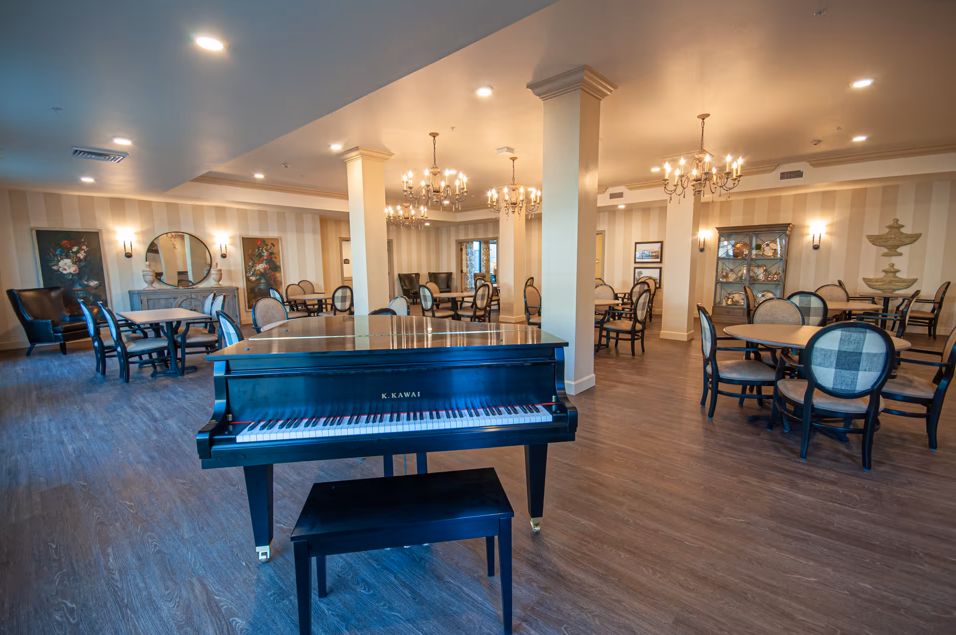 Spacious senior living lounge with grand piano, round tables, upholstered chairs, and chandeliers.