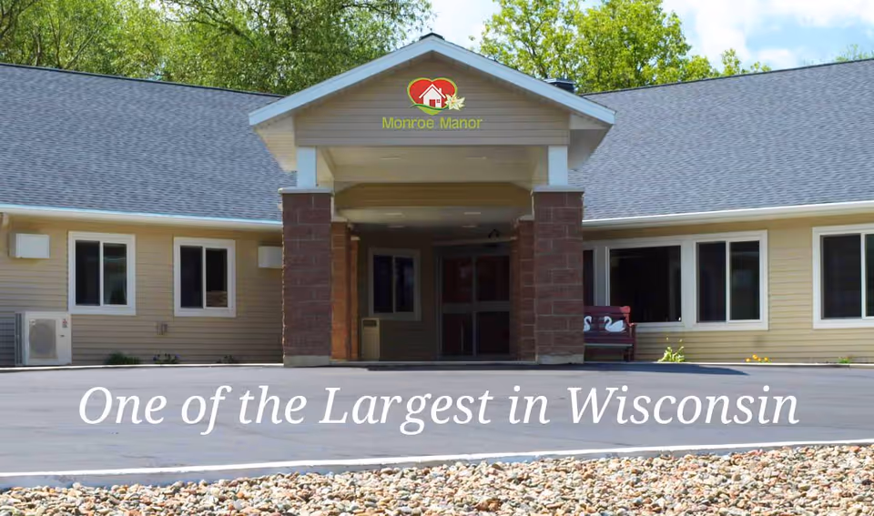 Front exterior view of Monroe Manor, a single-story building with beige siding, a gray roof, and a covered entrance supported by brick columns. There is a paved driveway and a rock bed in front of the building. The text 'One of the Largest in Wisconsin' is overlaid at the bottom of the image.