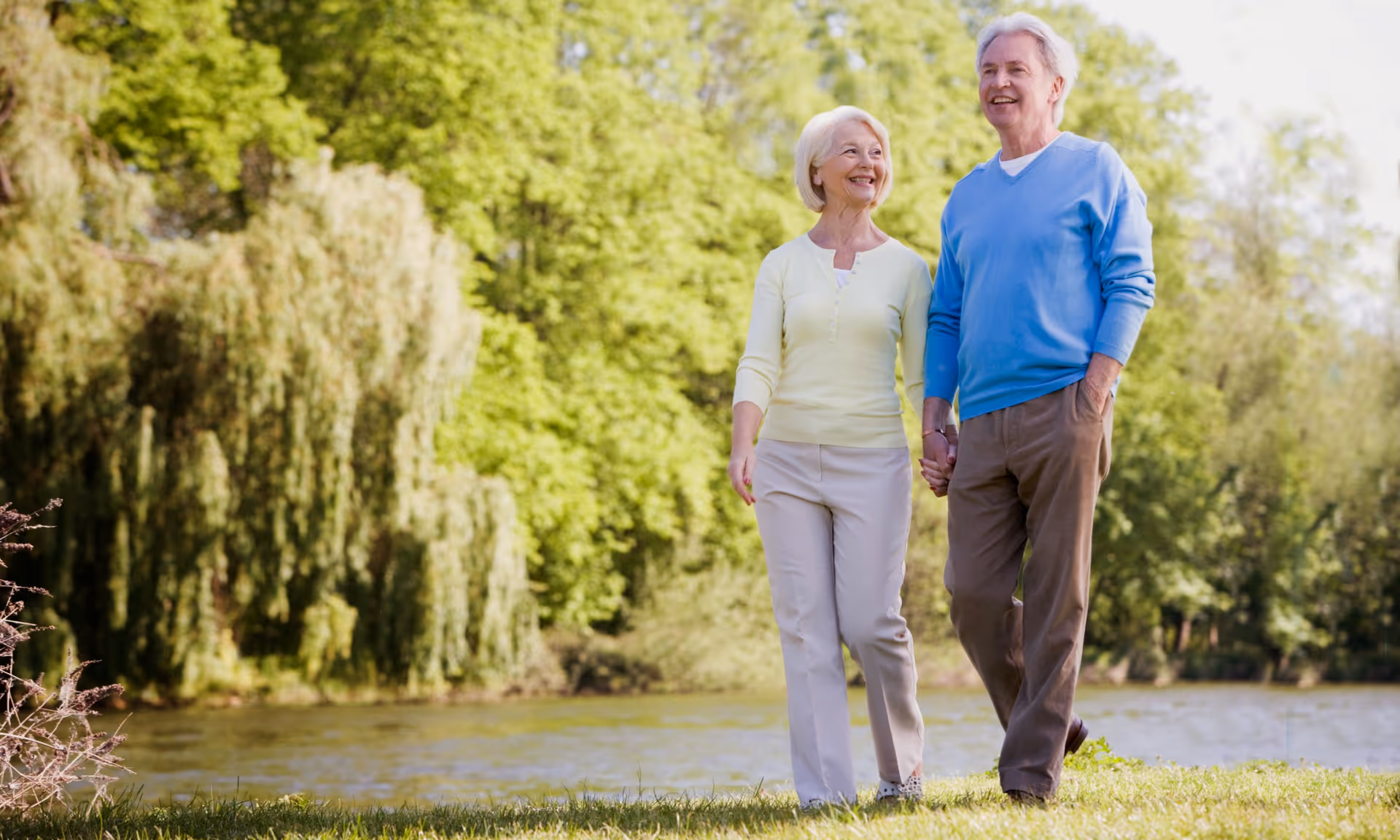 Two seniors walk hand-in-hand outdoors beside a lake surrounded by trees.