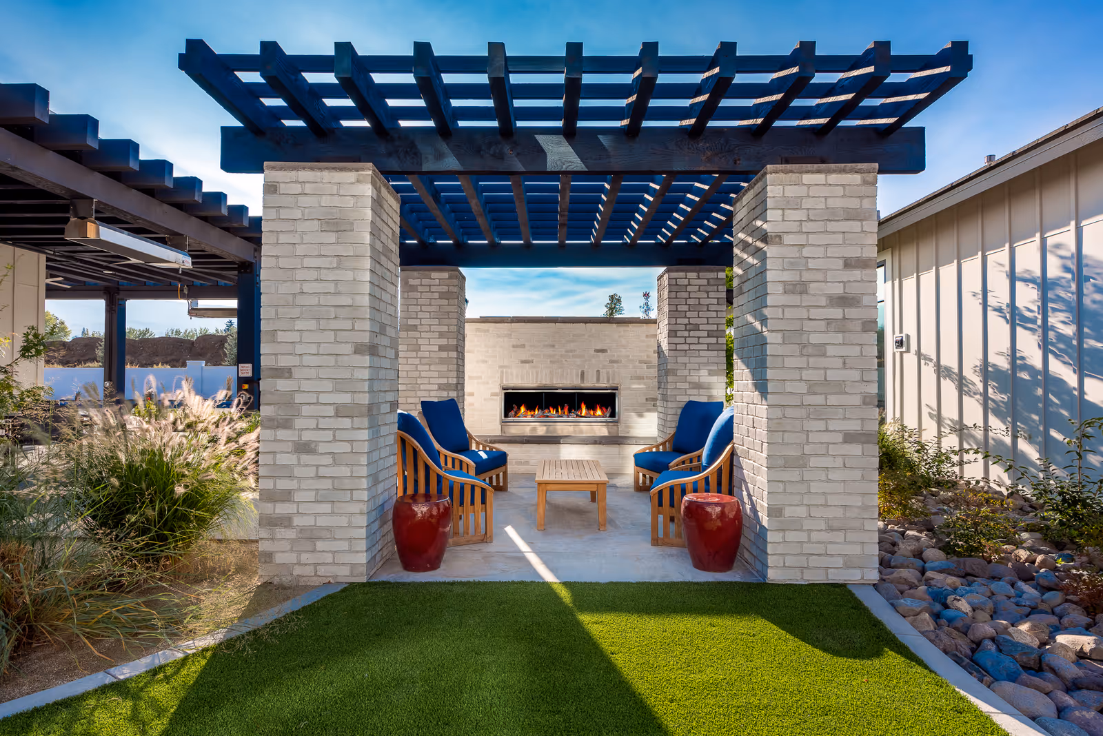 Outdoor seating area with four wooden chairs with blue cushions arranged around a small wooden table under a pergola. There is a modern outdoor fireplace built into a light brick wall at the back. The area is surrounded by greenery and landscaping with a clear blue sky overhead.