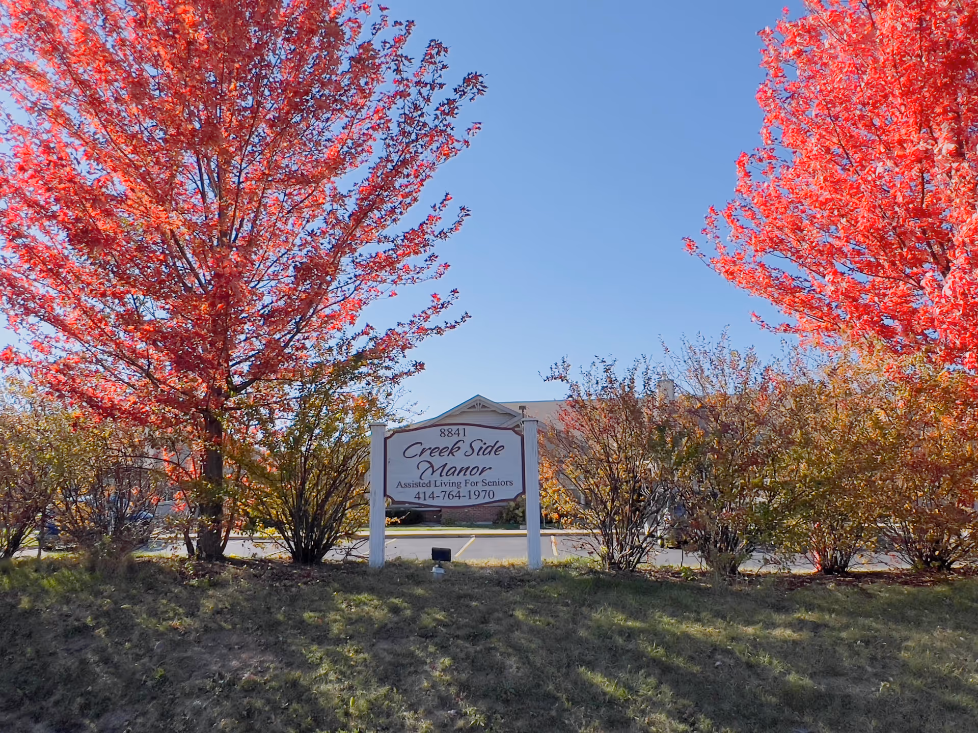 Outdoor view of Caring Alternatives Creek Side Manor sign surrounded by bushes and trees with bright red autumn leaves under a clear blue sky.