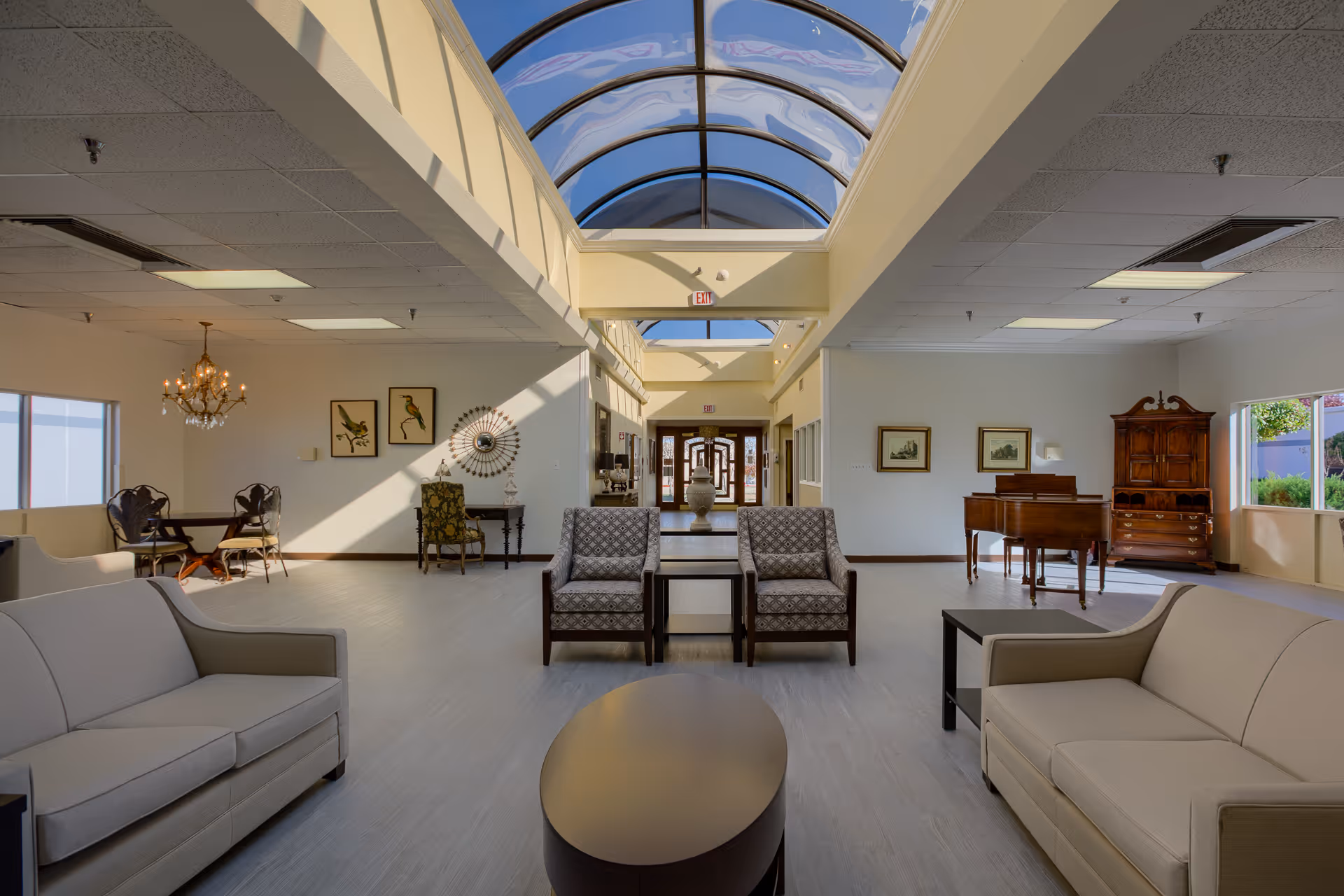 A spacious and well-lit common area with beige sofas, patterned armchairs, and a wooden coffee table. The room features a high ceiling with a large skylight, allowing natural light to fill the space. In the background, there is a wooden piano, framed artwork on the walls, and a small dining table with chairs under a chandelier. Large windows on the right side provide views of greenery outside.