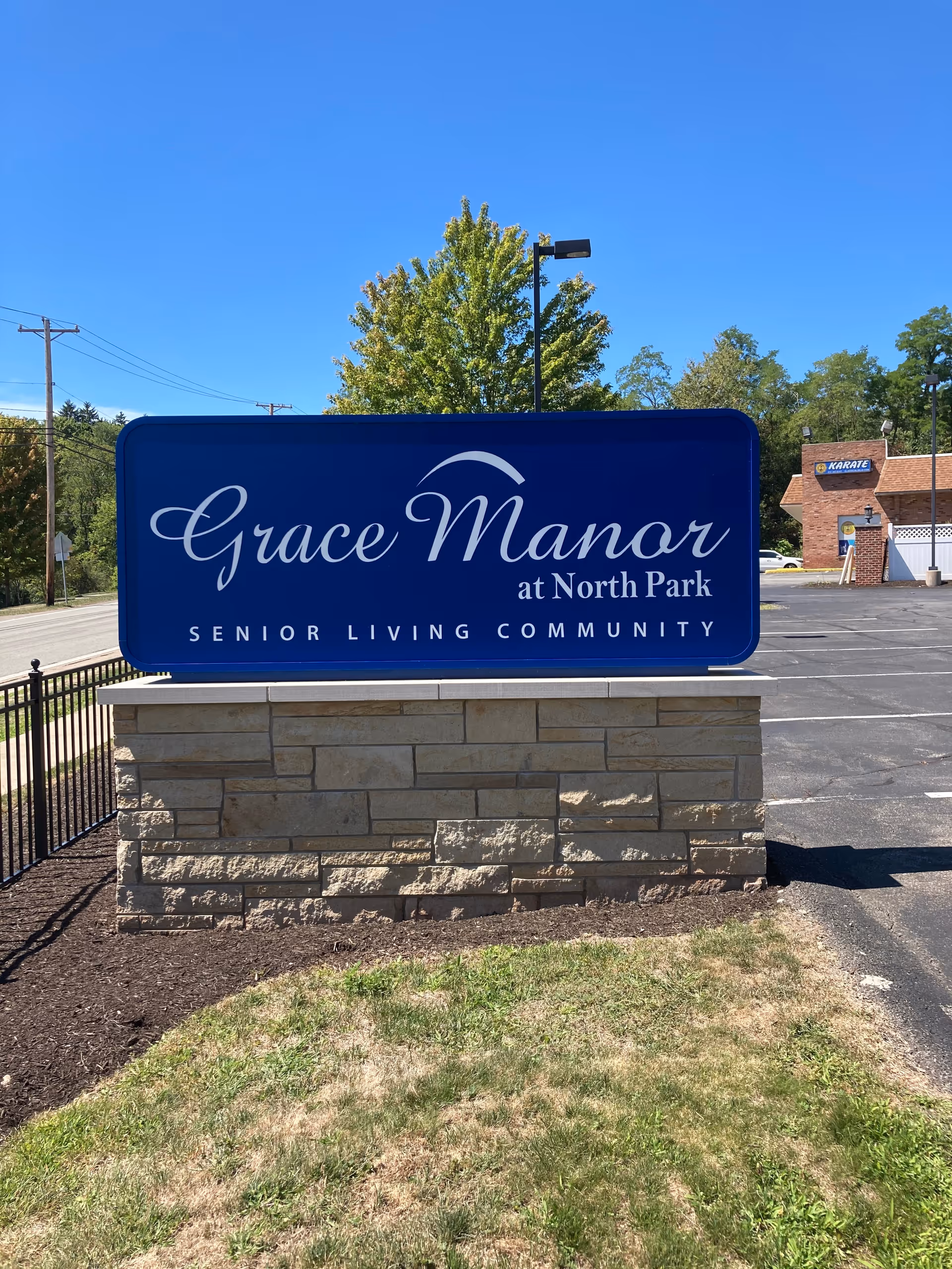 A large blue sign with white text that reads 'Grace Manor at North Park Senior Living Community' mounted on a stone base, with a parking lot and trees in the background under a clear blue sky.