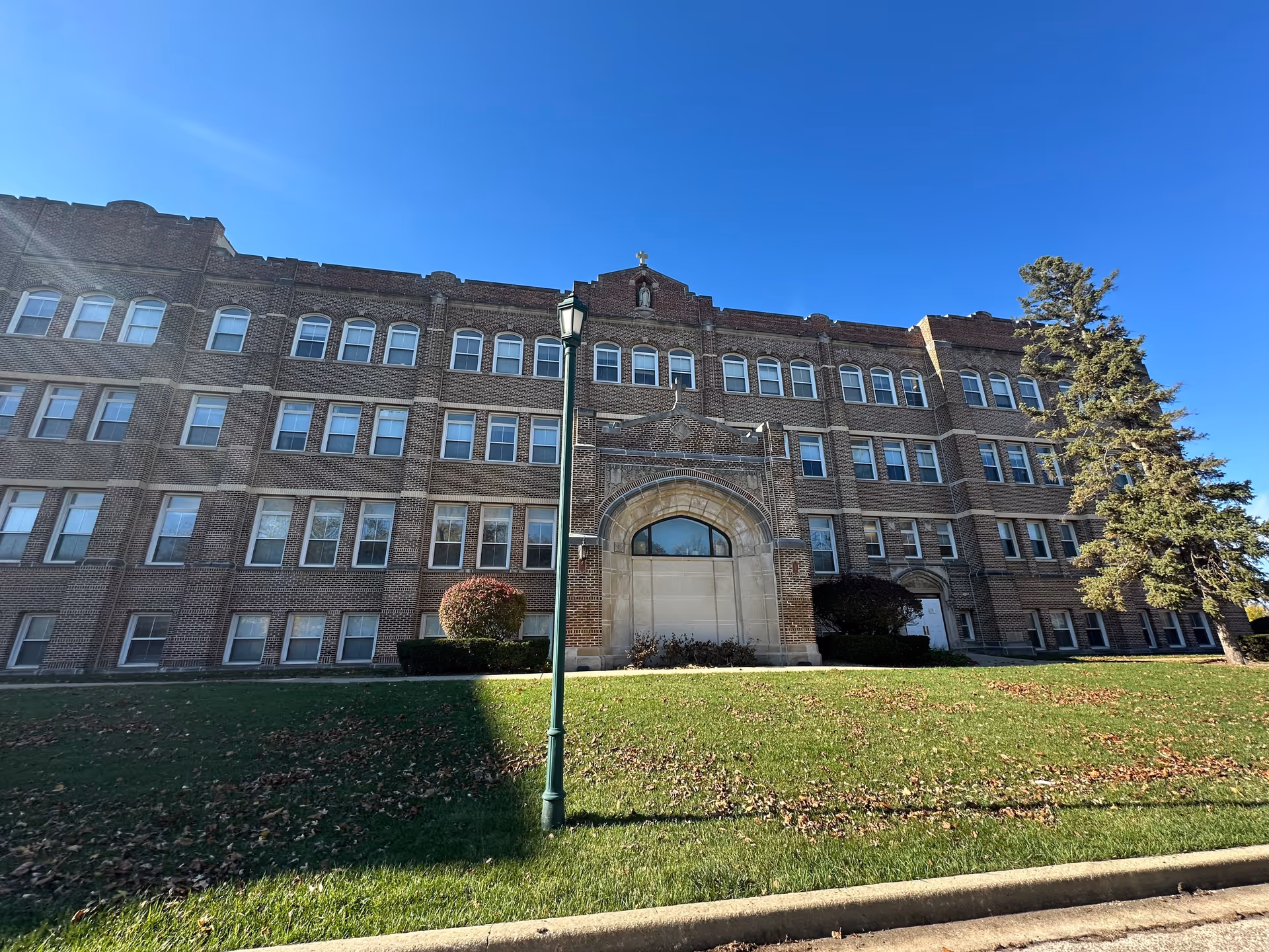 Exterior view of a large, multi-story brick building with many windows, a decorative arched entrance, a green lawn with scattered leaves, a tall street lamp, and a tree on the right side under a clear blue sky.