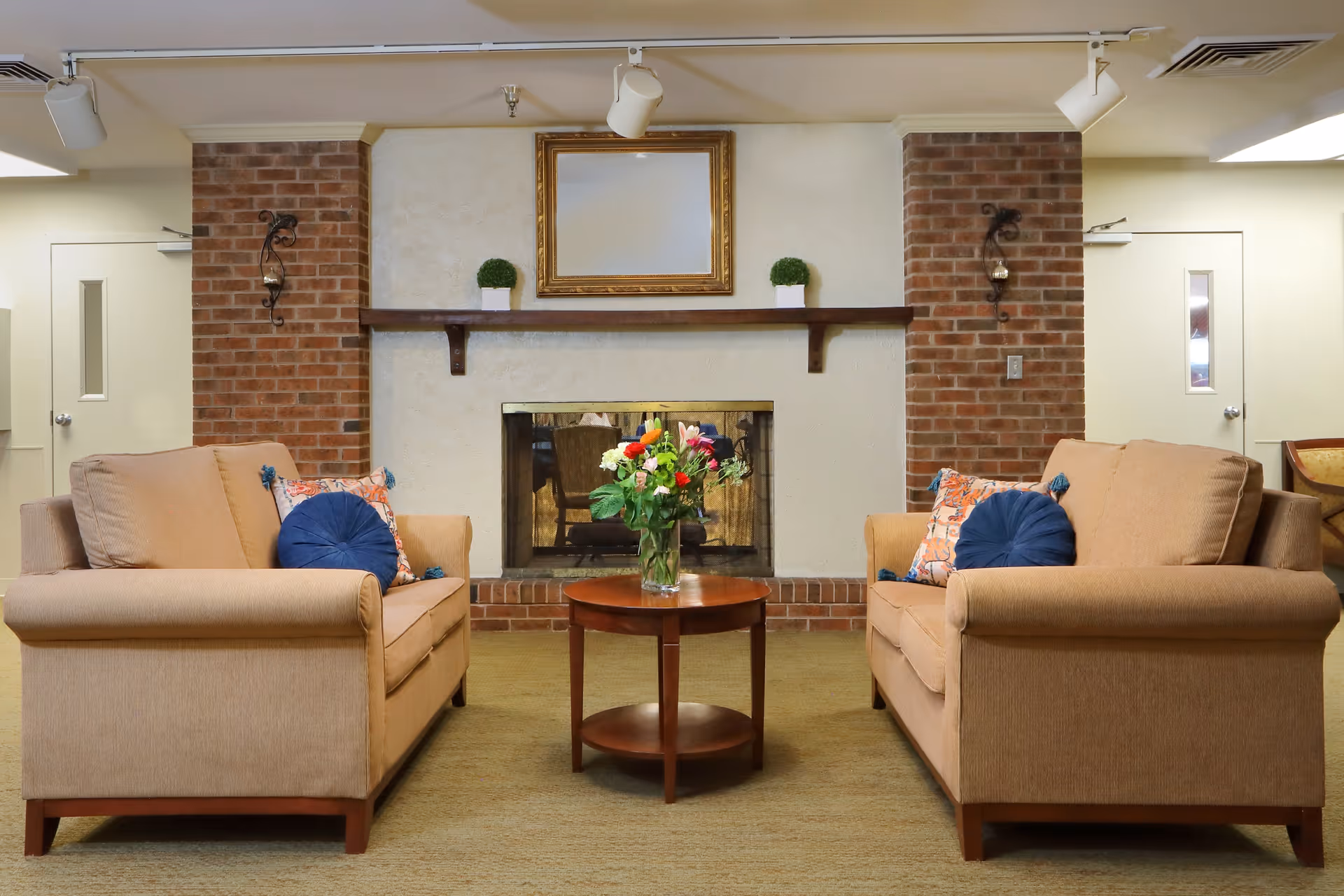 A cozy living room area with two beige sofas facing each other, each adorned with blue and patterned throw pillows. Between the sofas is a round wooden coffee table with a vase of colorful flowers. The background features a brick fireplace with a wooden mantel holding two small potted plants and a large empty picture frame above it. The walls are light-colored, and there are two doors on either side of the fireplace.