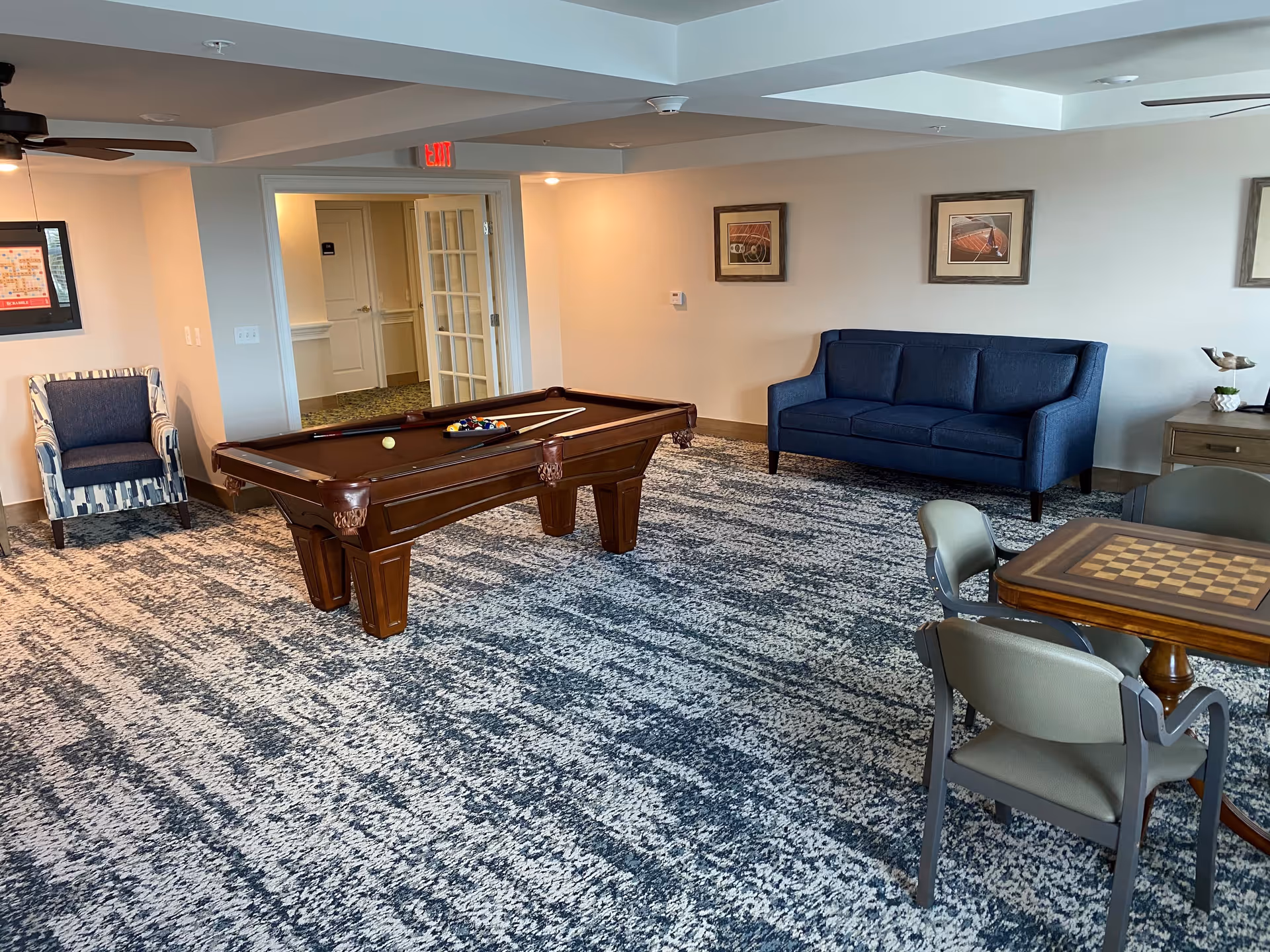 A recreational room with a pool table in the center, a blue couch against the wall, a patterned armchair, and a table with a chessboard and chairs. The room has a patterned carpet, framed pictures on the walls, and a ceiling fan.