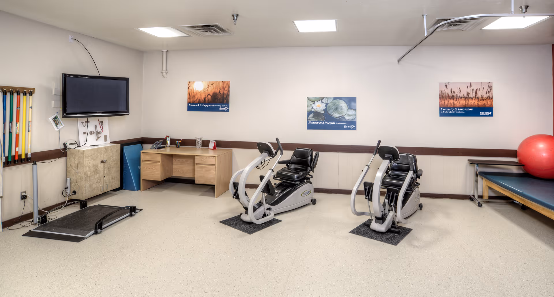 A rehabilitation room with exercise equipment including two recumbent exercise bikes, a treadmill, a desk with office supplies, a large red exercise ball on a padded table, and a wall-mounted TV. The walls have three framed motivational posters.