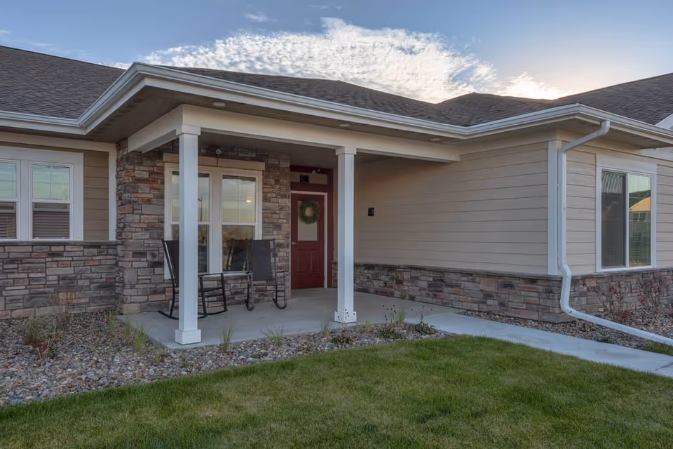 A single-story senior living facility with a covered front porch, red door, stone accents, and two chairs on the entry patio.