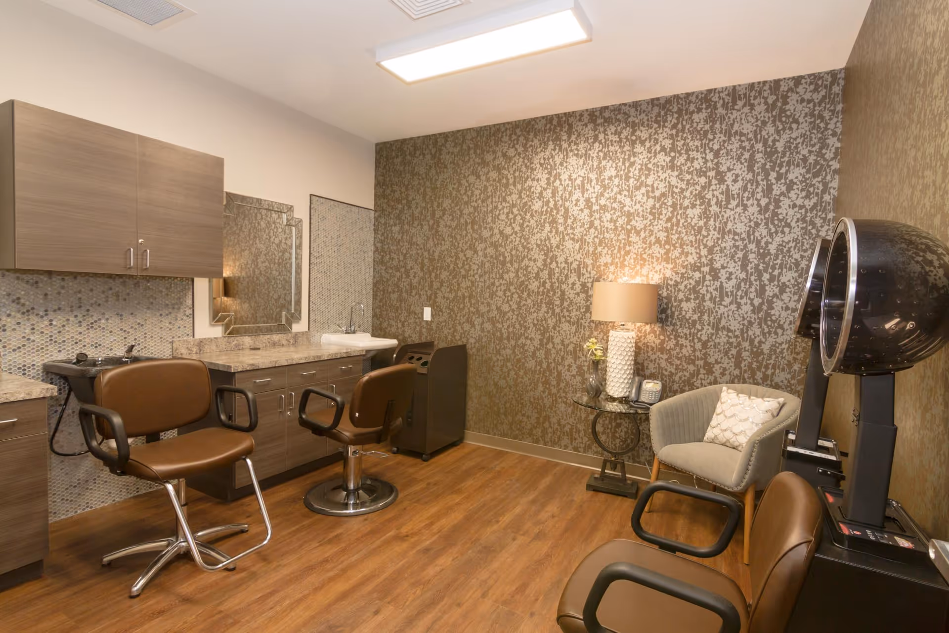 Interior of a salon room with two brown salon chairs in front of a counter with a mirror and sink. The room has wood flooring, patterned wallpaper, a small round glass table with a lamp and a phone, a gray armchair with a decorative pillow, and a black hair dryer hood.