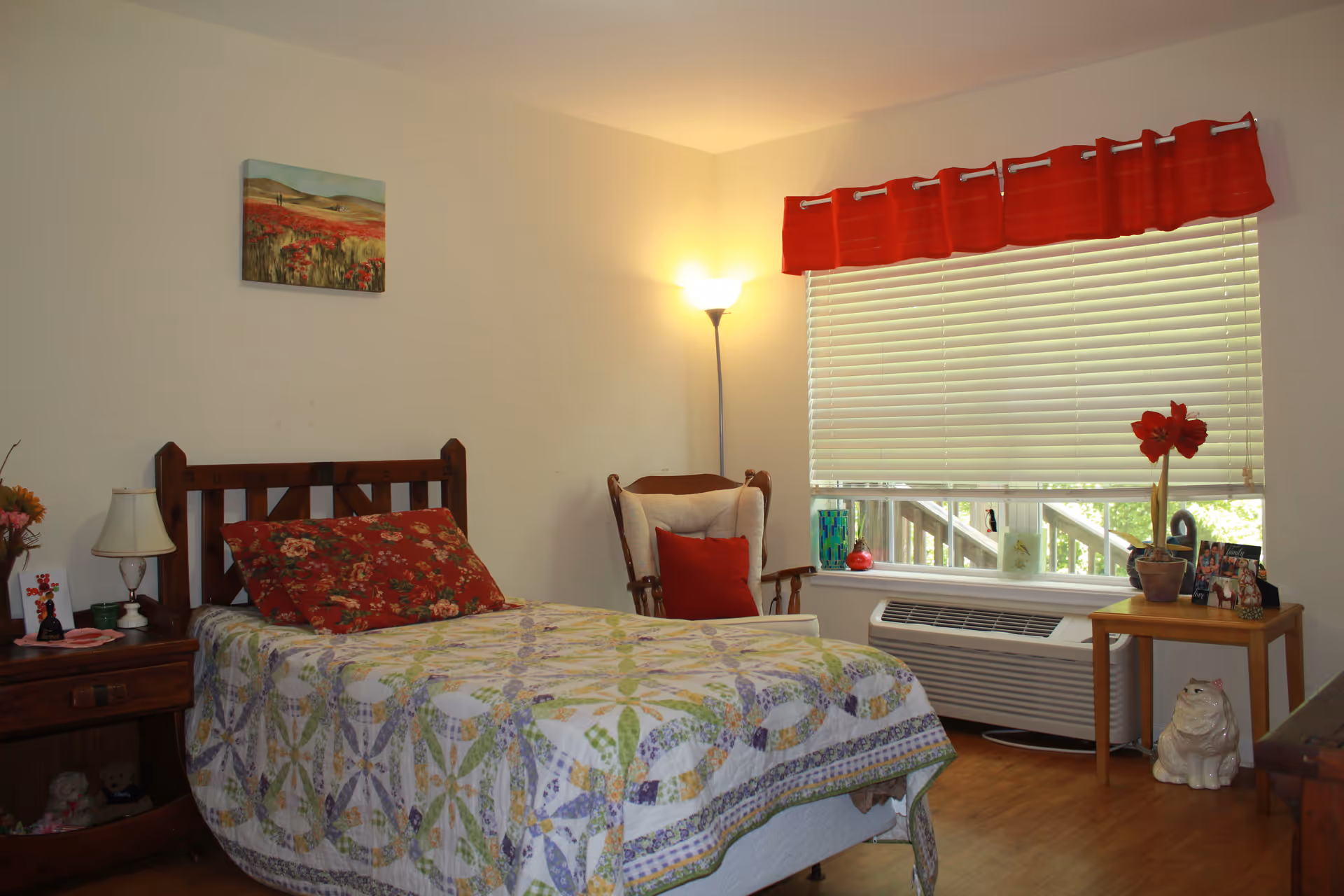 A cozy bedroom with a wooden bed frame, floral bedding, and a large red floral pillow. There is a wooden nightstand with a lamp and decorative items on the left side of the bed. A cushioned armchair with a red pillow is placed near a window with white blinds and a red valance. A small wooden table with a potted plant and framed photos is next to the window. The room has light-colored walls, a floor lamp in the corner, and wooden flooring.