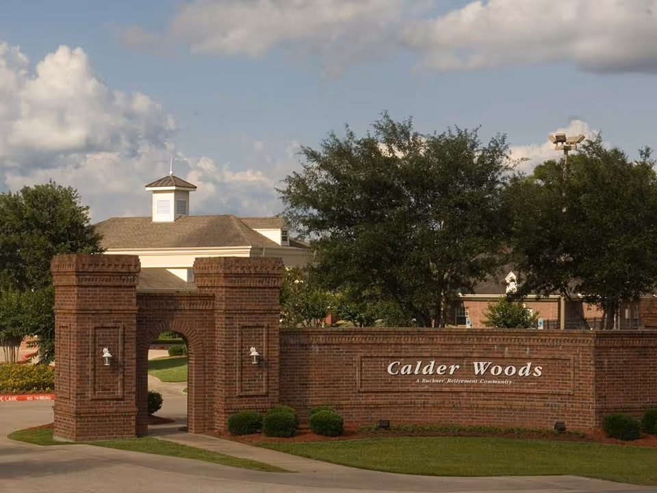 Entrance to Calder Woods retirement community featuring a brick archway and a brick wall with the community's name displayed. Trees and a building with a cupola are visible in the background under a partly cloudy sky.