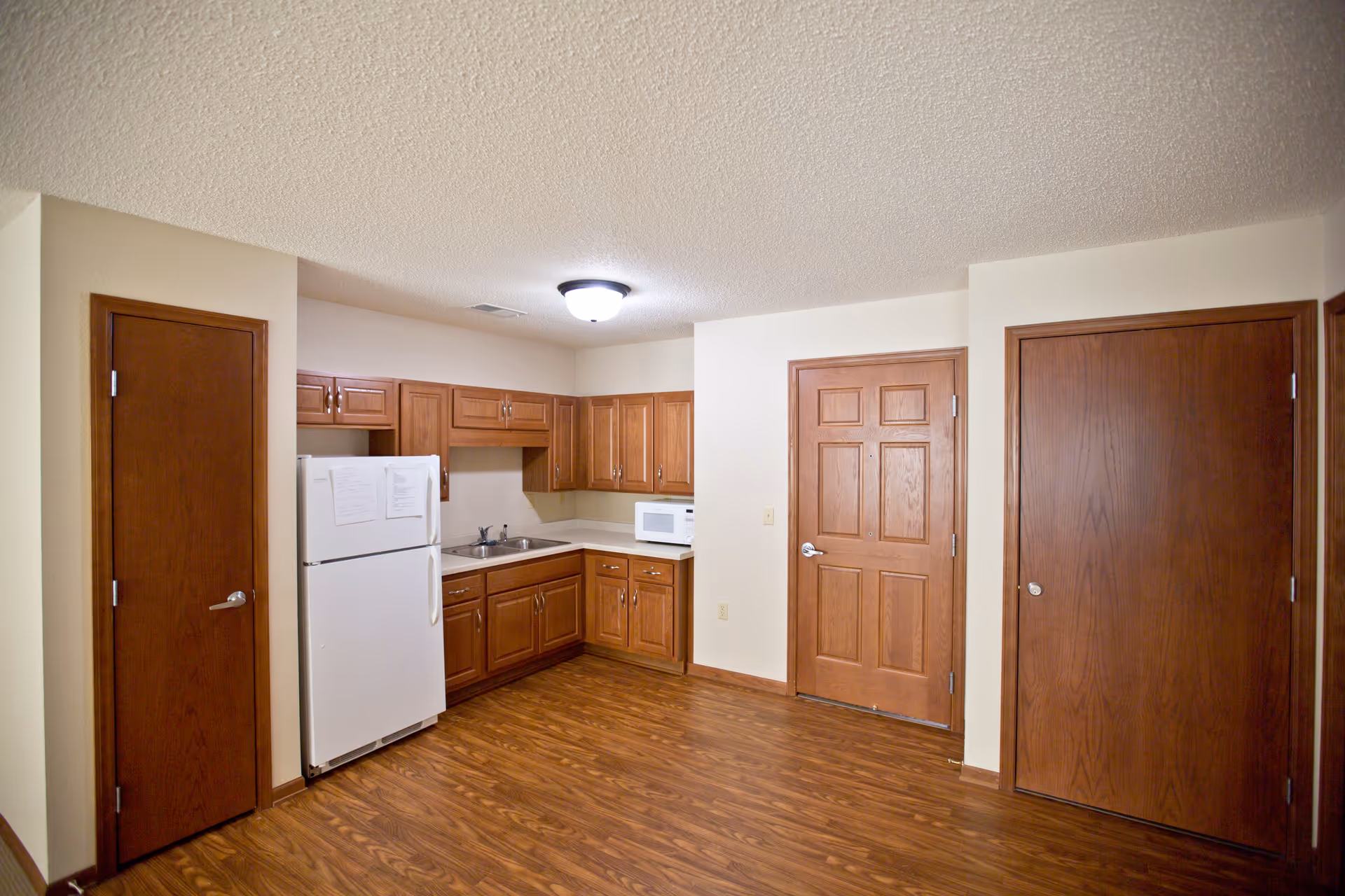 Interior view of a small kitchen area with wooden cabinets, a white refrigerator, a microwave, a sink, and three wooden doors. The floor is wood laminate and the ceiling has a textured finish with a ceiling light fixture.