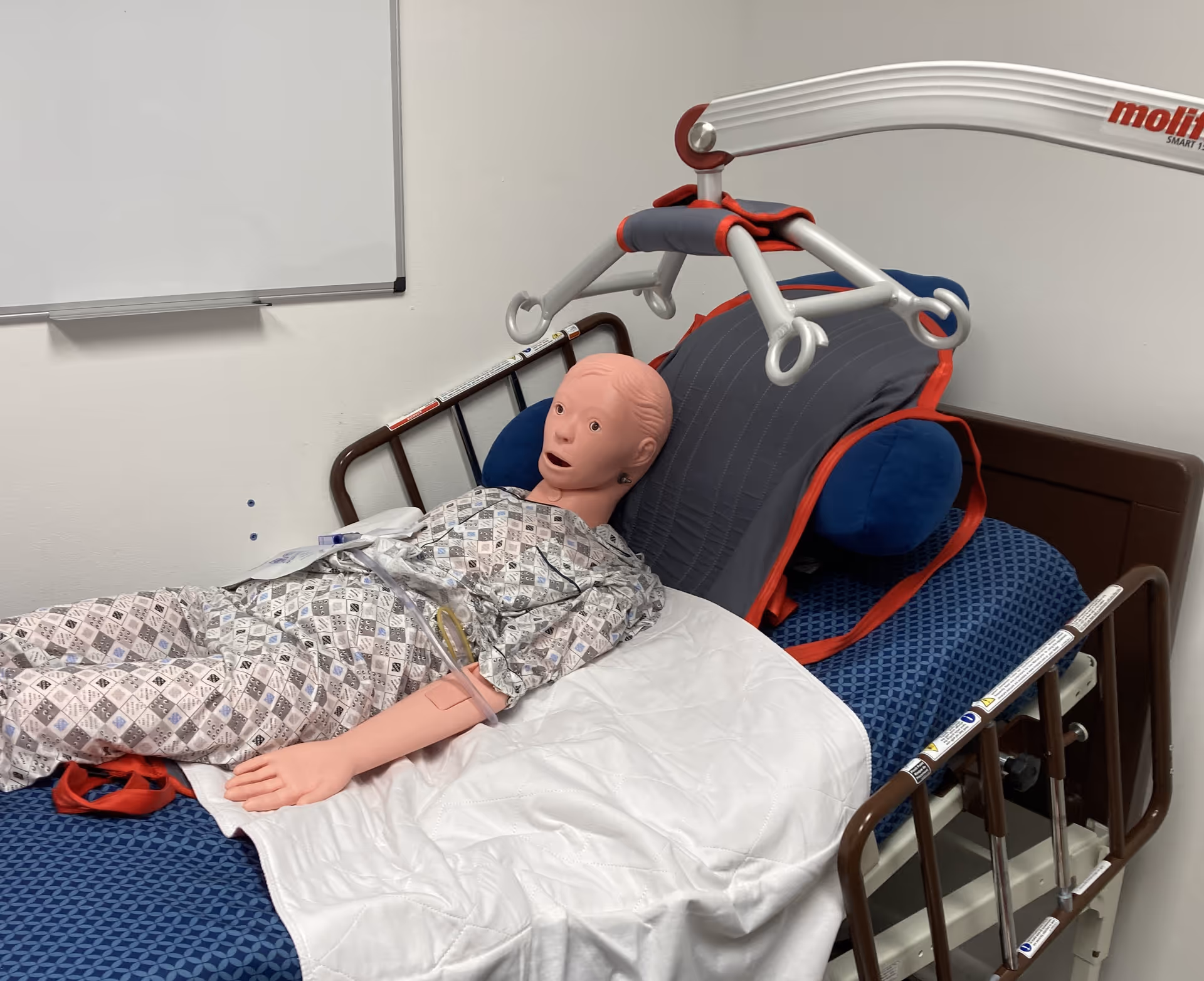 Medical training mannequin lying in hospital bed with overhead lift device indoors.