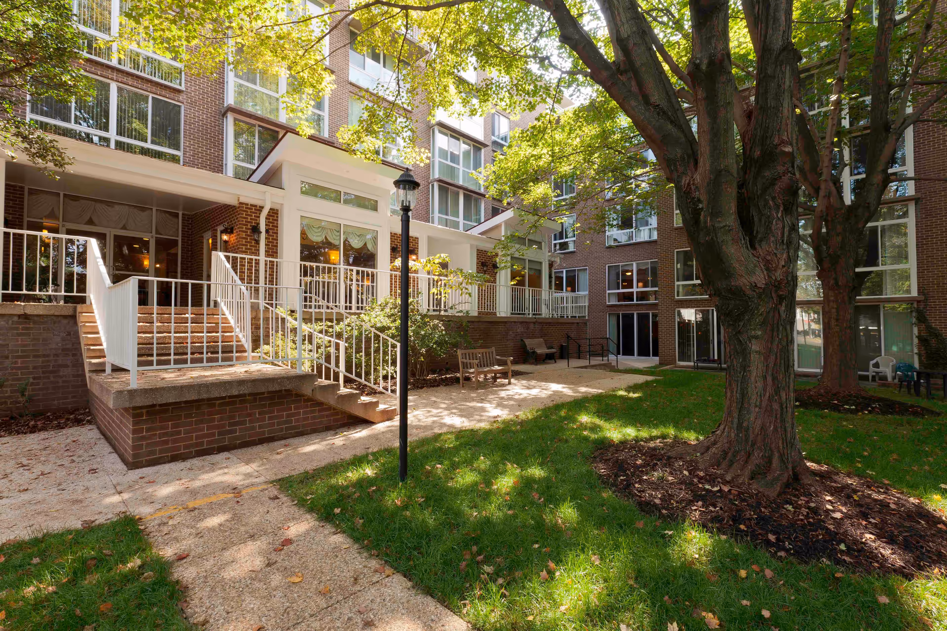 Outdoor courtyard area of a senior living facility with large trees, green grass, benches, and a brick building with multiple windows and white railings. Sunlight filters through the tree leaves, casting shadows on the ground.