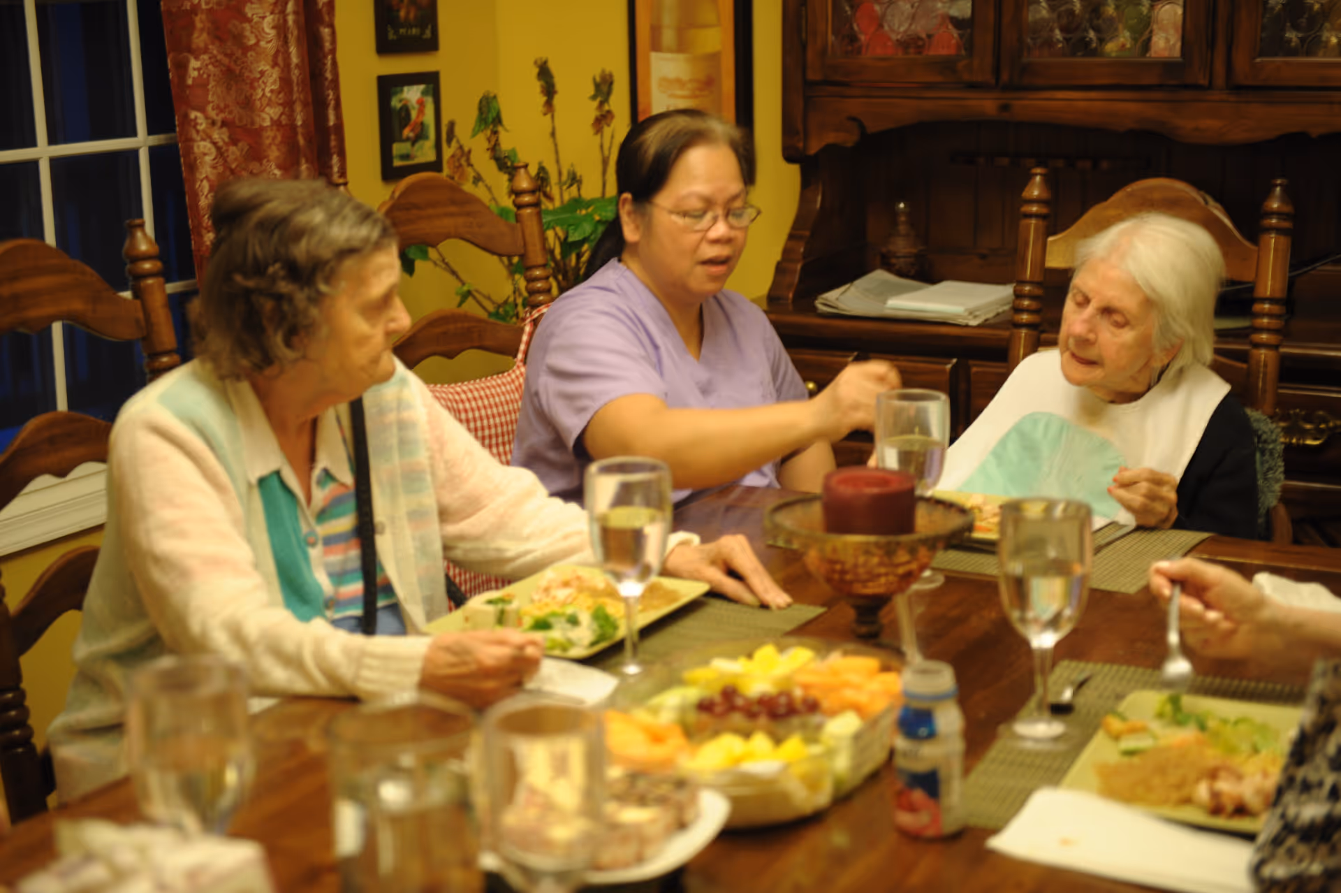 Three elderly women and a caregiver sitting around a wooden dining table having a meal together. The table is set with plates of food, glasses of water, and a centerpiece with a red candle. The room has warm yellow walls and wooden furniture.