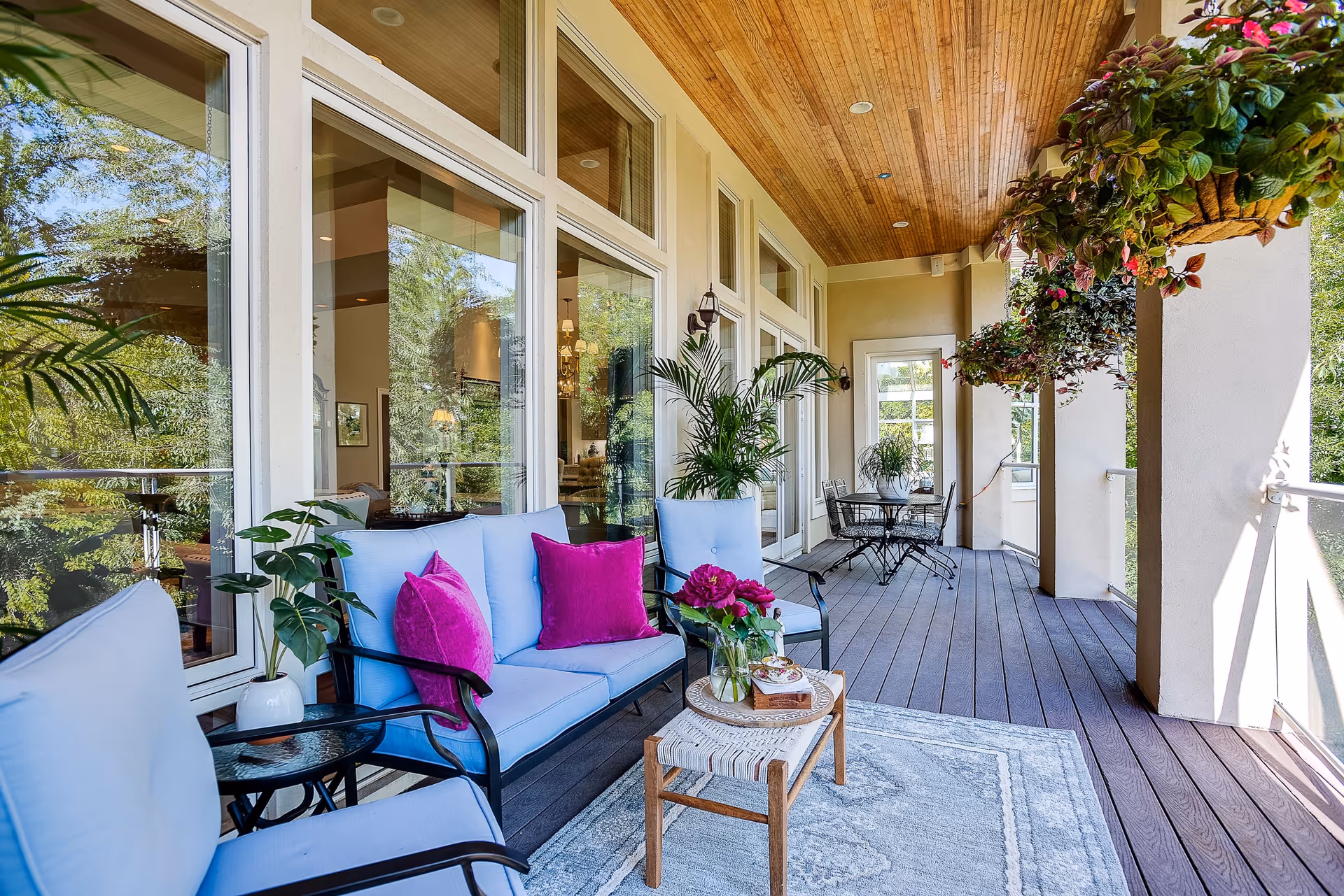 A spacious outdoor balcony area with light blue cushioned seating, purple throw pillows, a small wooden table with flowers, and hanging plants. The balcony has a wooden ceiling, large windows reflecting greenery, and a small dining table with chairs at the far end.