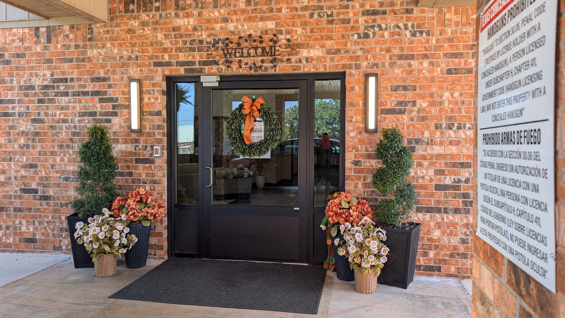 Brick building entrance with glass doors, wreath, and potted plants flanking doorway.