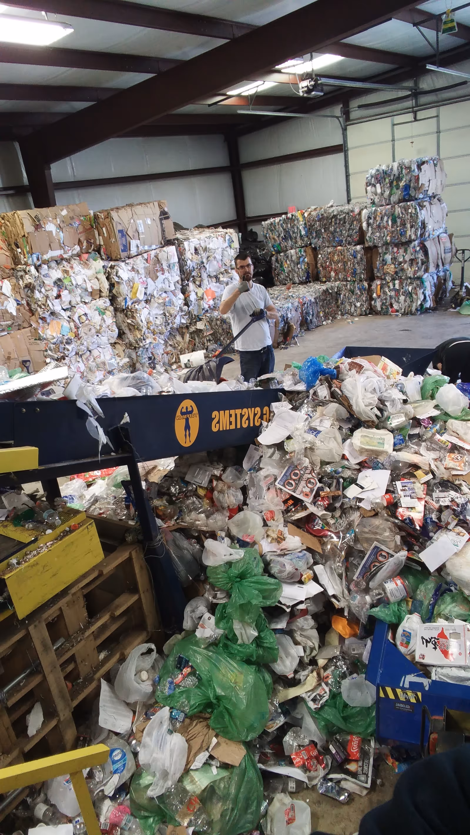 Warehouse interior overflowing with stacked recycling bales and a worker amid cluttered piles.