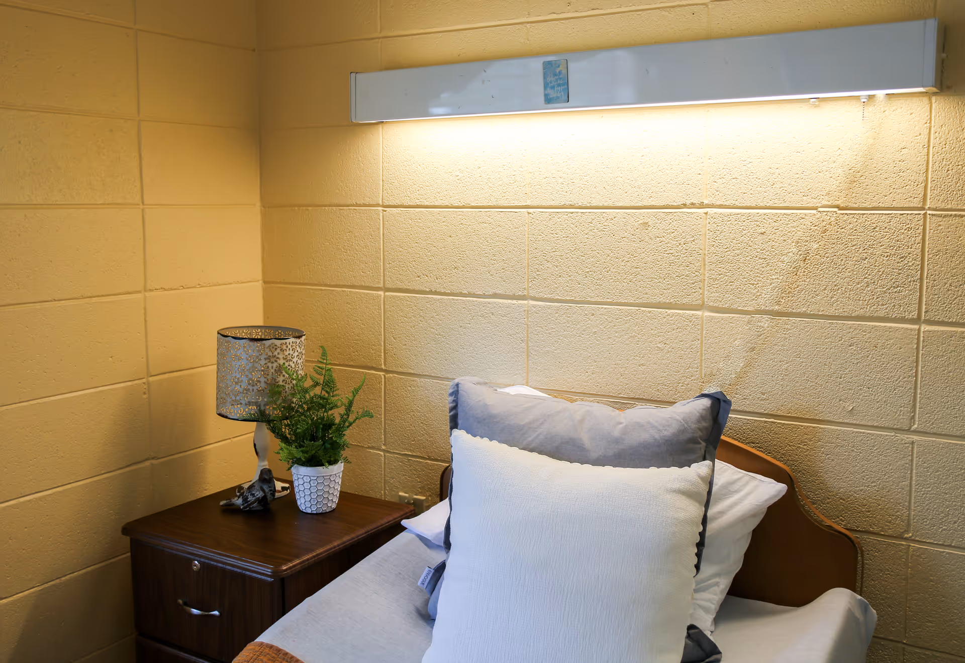 A small bed with white and gray pillows against a yellow brick wall. Next to the bed is a wooden nightstand with a decorative lamp and a potted green plant. A long light fixture is mounted on the wall above the bed.