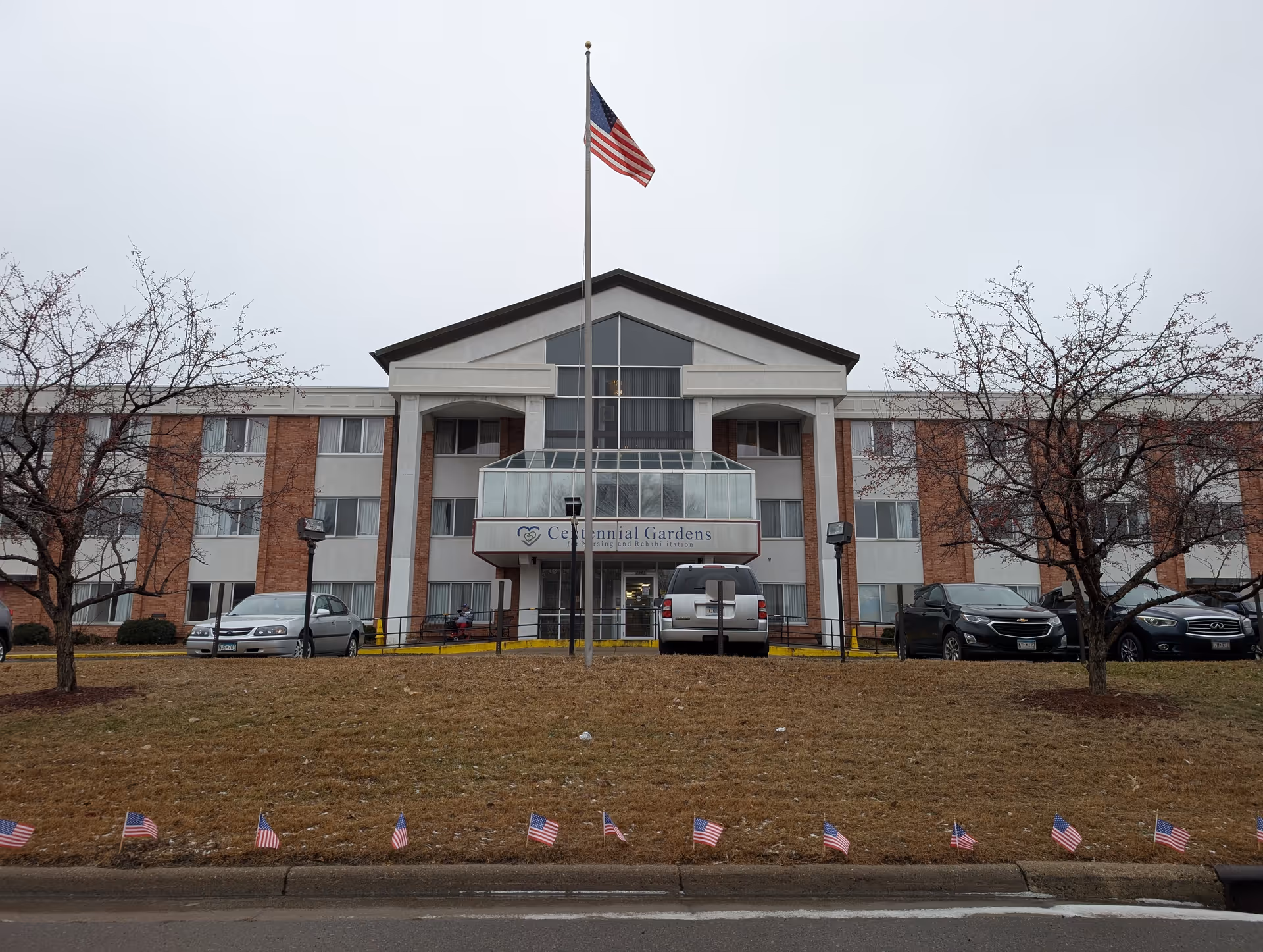 Front exterior view of Centennial Gardens for Nursing and Rehabilitation, a three-story building with a central glass entrance and an American flag flying on a flagpole in front. Several cars are parked near the entrance, and small American flags line the grass along the sidewalk.