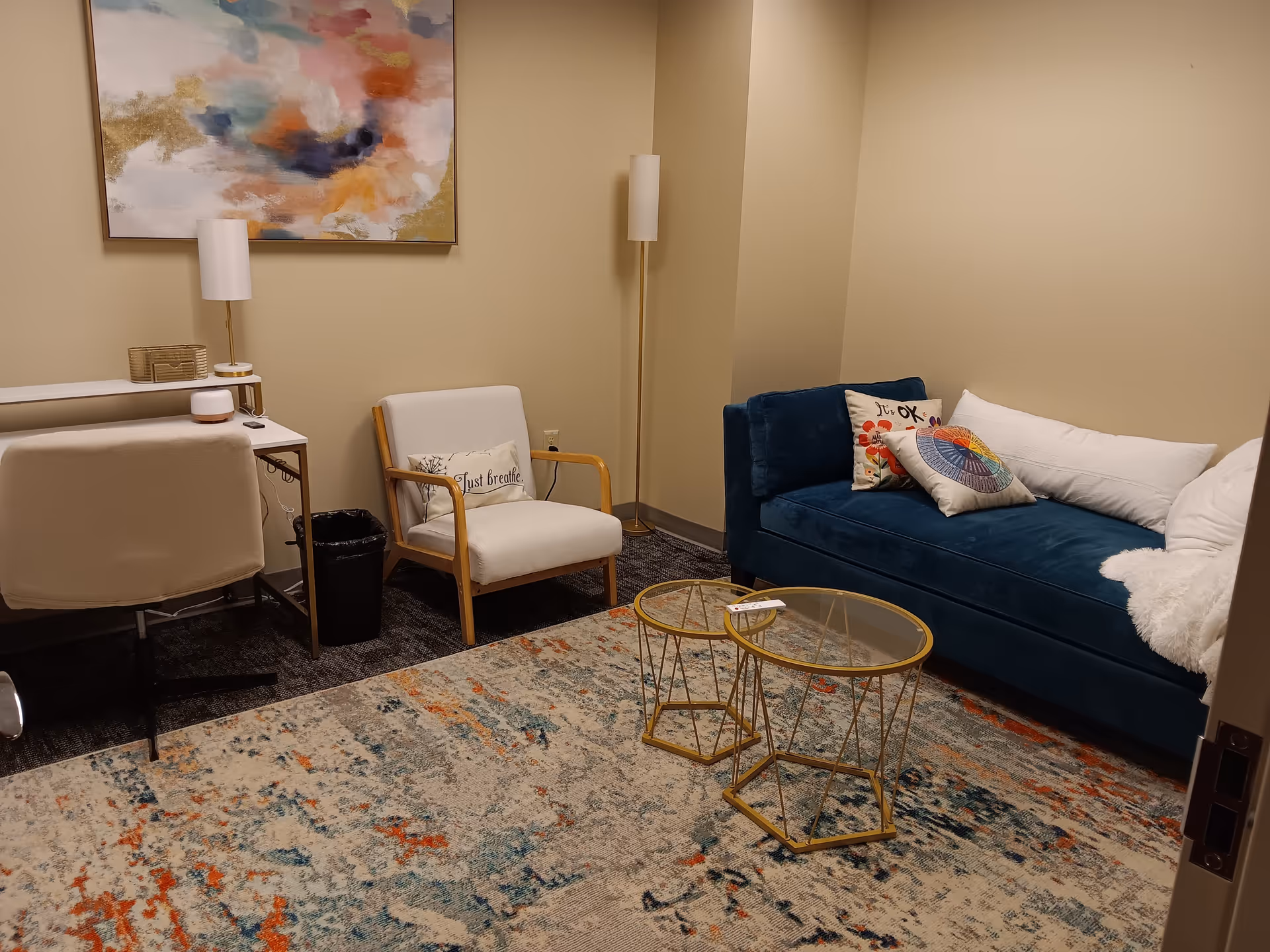 A cozy lounge with a blue sofa, white chair, abstract painting, and glass coffee tables.