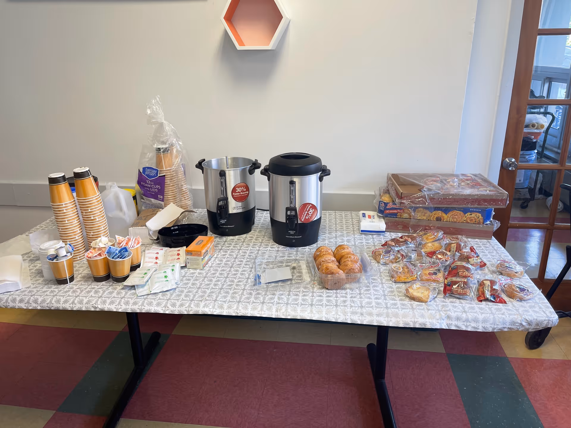 A snack and coffee service table with pastries, cups, and urns in a communal room.