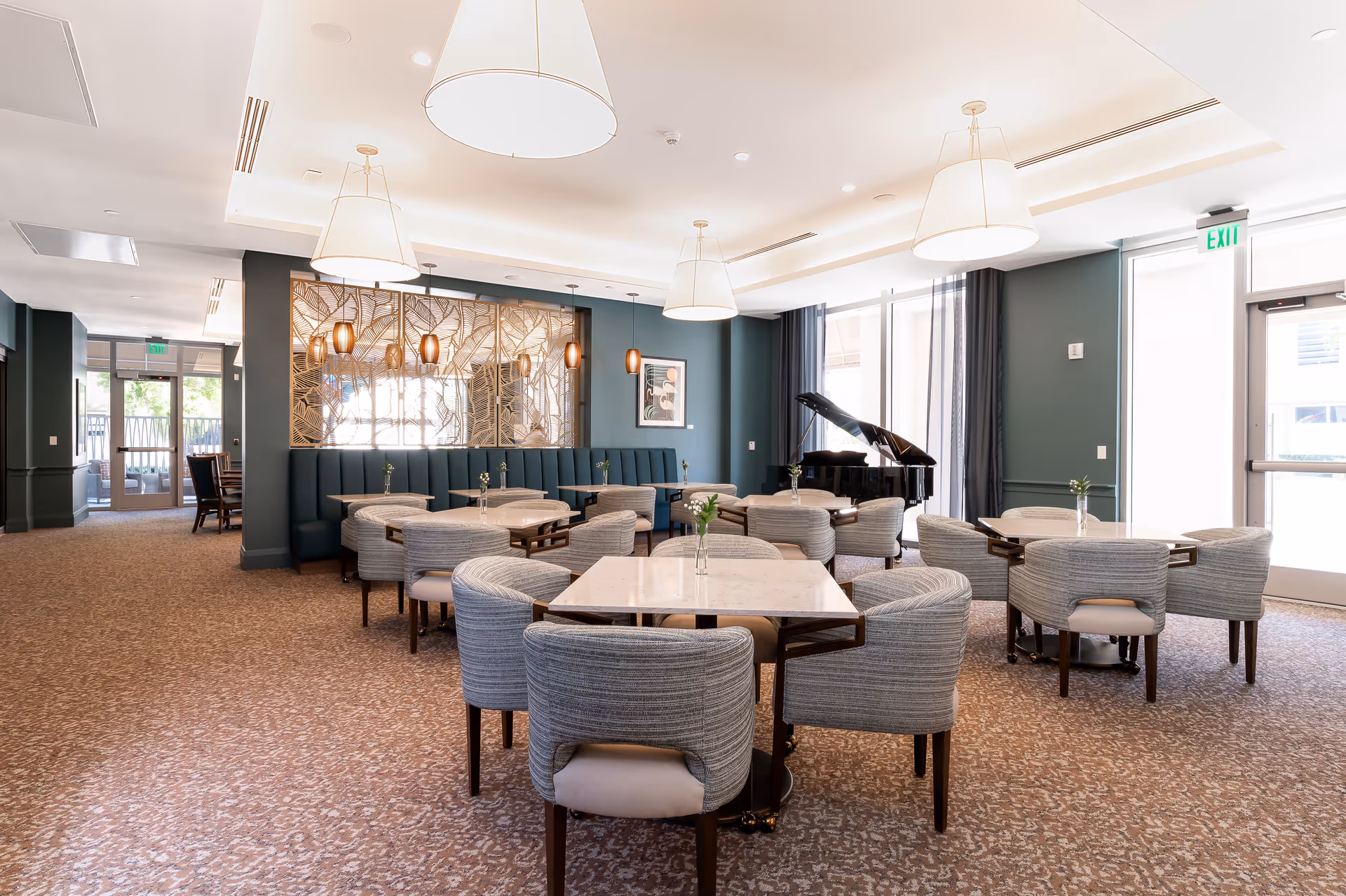 A spacious, well-lit dining area with multiple round tables surrounded by cushioned chairs. The room features a grand piano near large windows with sheer curtains, modern pendant lights hanging from the ceiling, and decorative wall panels with warm lighting. The carpet has a subtle pattern, and there is an exit door on the right side.