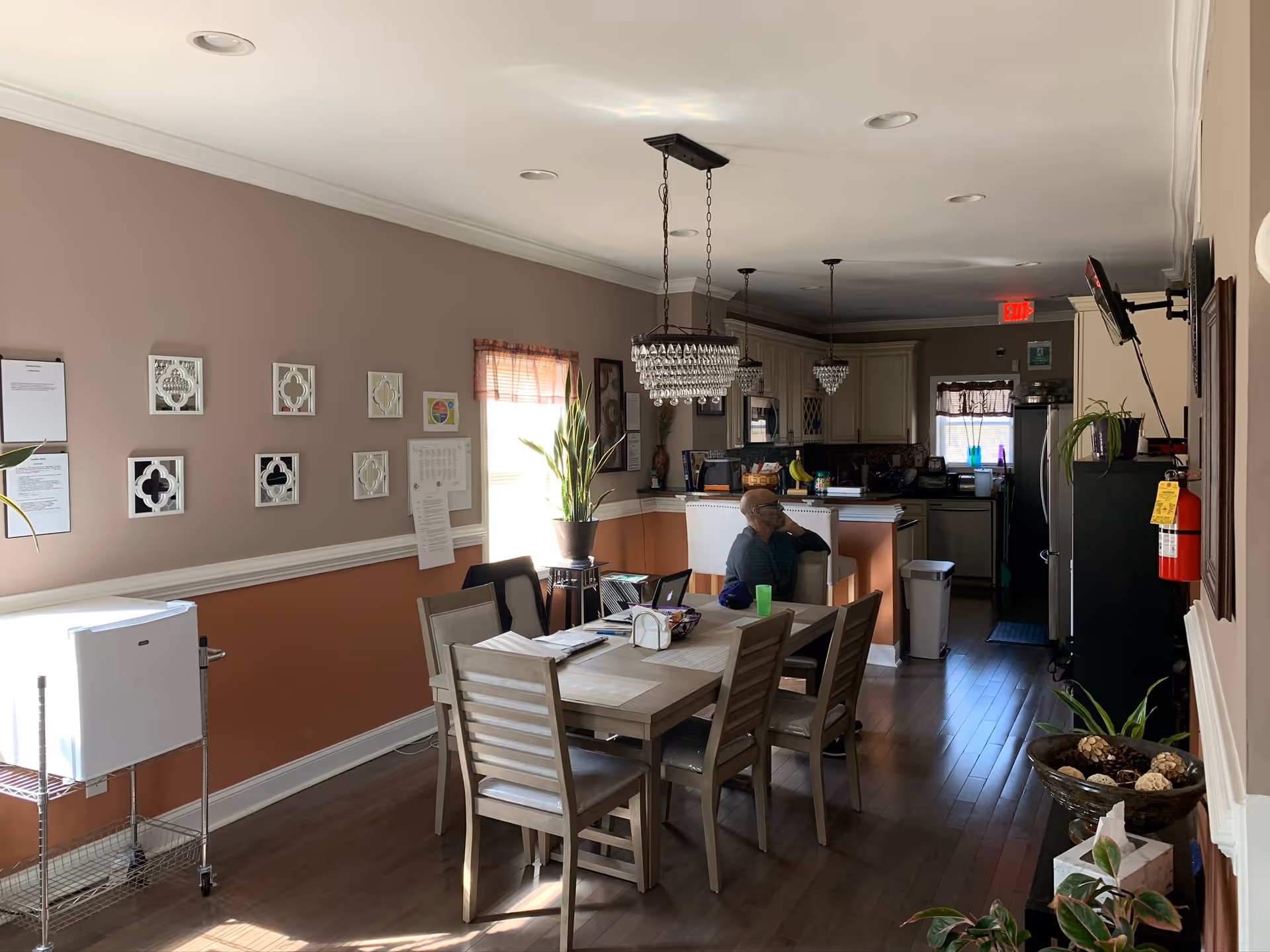 Interior view of a dining area and kitchen in an assisted living facility. The dining area has a wooden table with six chairs, some papers and a green cup on the table. A man is sitting at the table looking towards the kitchen. The kitchen has cream-colored cabinets, a refrigerator, and a counter with bar stools. The walls are painted in two tones, with decorative mirrors and framed papers on one side. There are plants and a fire extinguisher visible, and the room is lit by natural light from a window and ceiling lights.