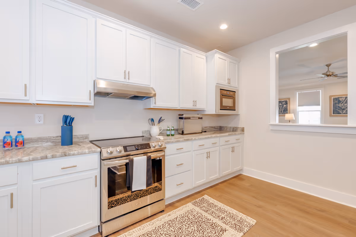 White kitchen with stainless stove, microwave, quartz counters, patterned rug, and light wood flooring.