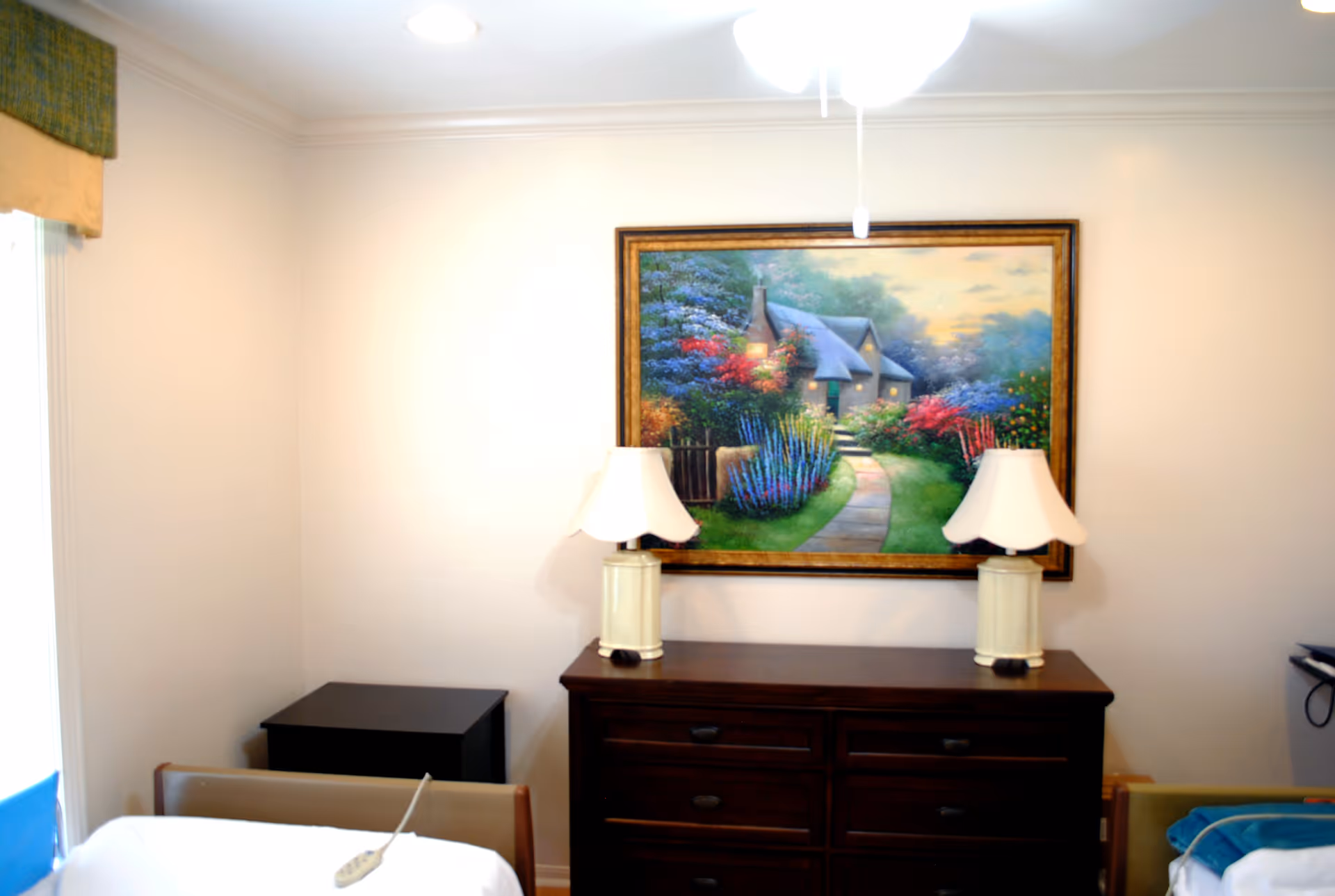 A bedroom with a bed, a dark dresser, two lamps, and a framed garden painting above.