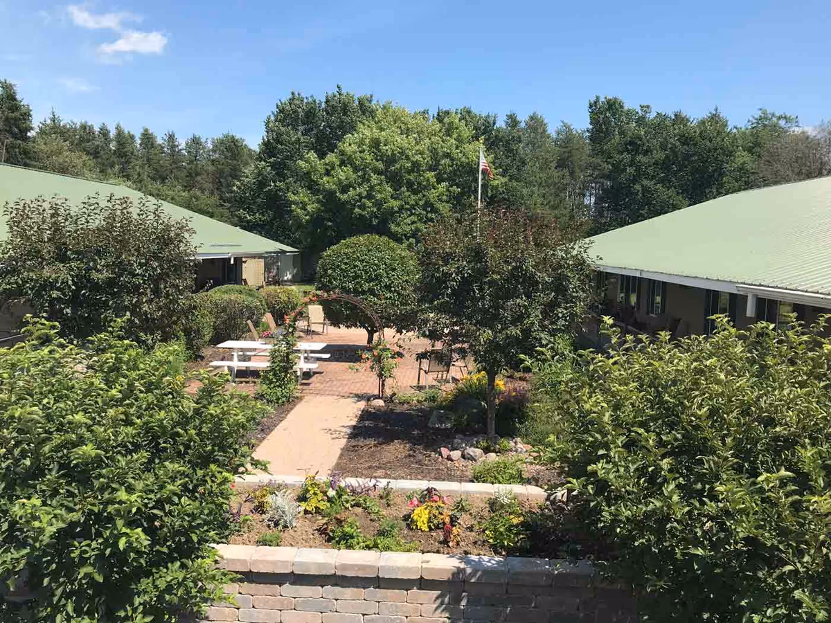 Outdoor courtyard area at Cedar Creek Manor with green trees, bushes, a brick-paved patio, white picnic tables, chairs, and a metal archway with climbing plants under a clear blue sky.