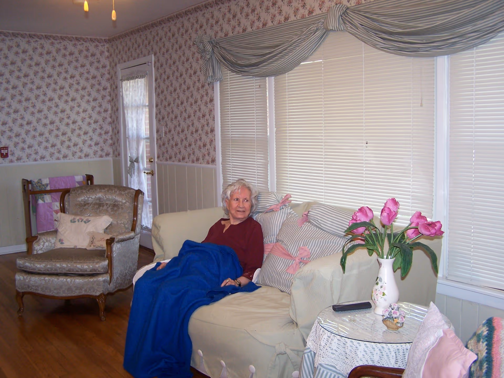 An older woman sits on a couch covered by a blue blanket in a floral-patterned living room with an armchair and a side table holding a vase of pink flowers.