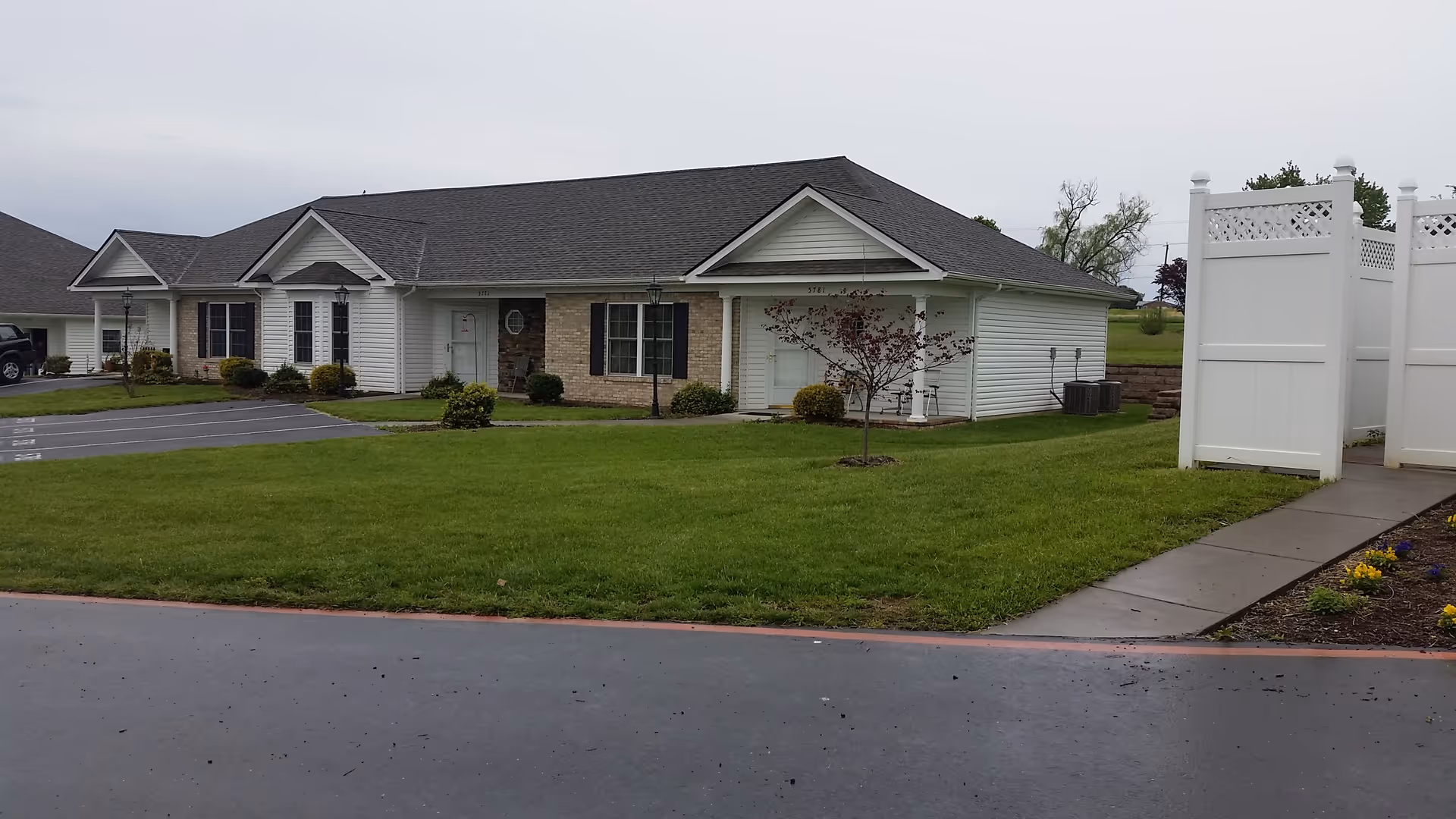 Single-story residential building with white siding and brick accents, surrounded by a well-maintained lawn and a paved driveway. There is a white privacy fence on the right side and a few small trees and shrubs in the yard under an overcast sky.