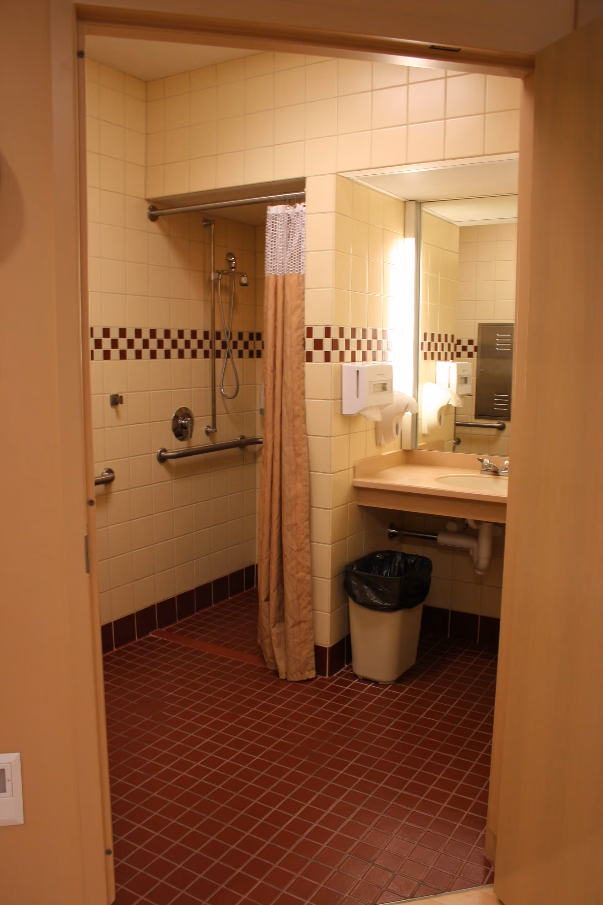 Accessible bathroom with beige tiles, a shower, sink, mirror, and trash can.