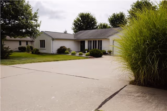 Single-story light-colored ranch-style building with a wide driveway, lawn and shrubs under a cloudy sky.