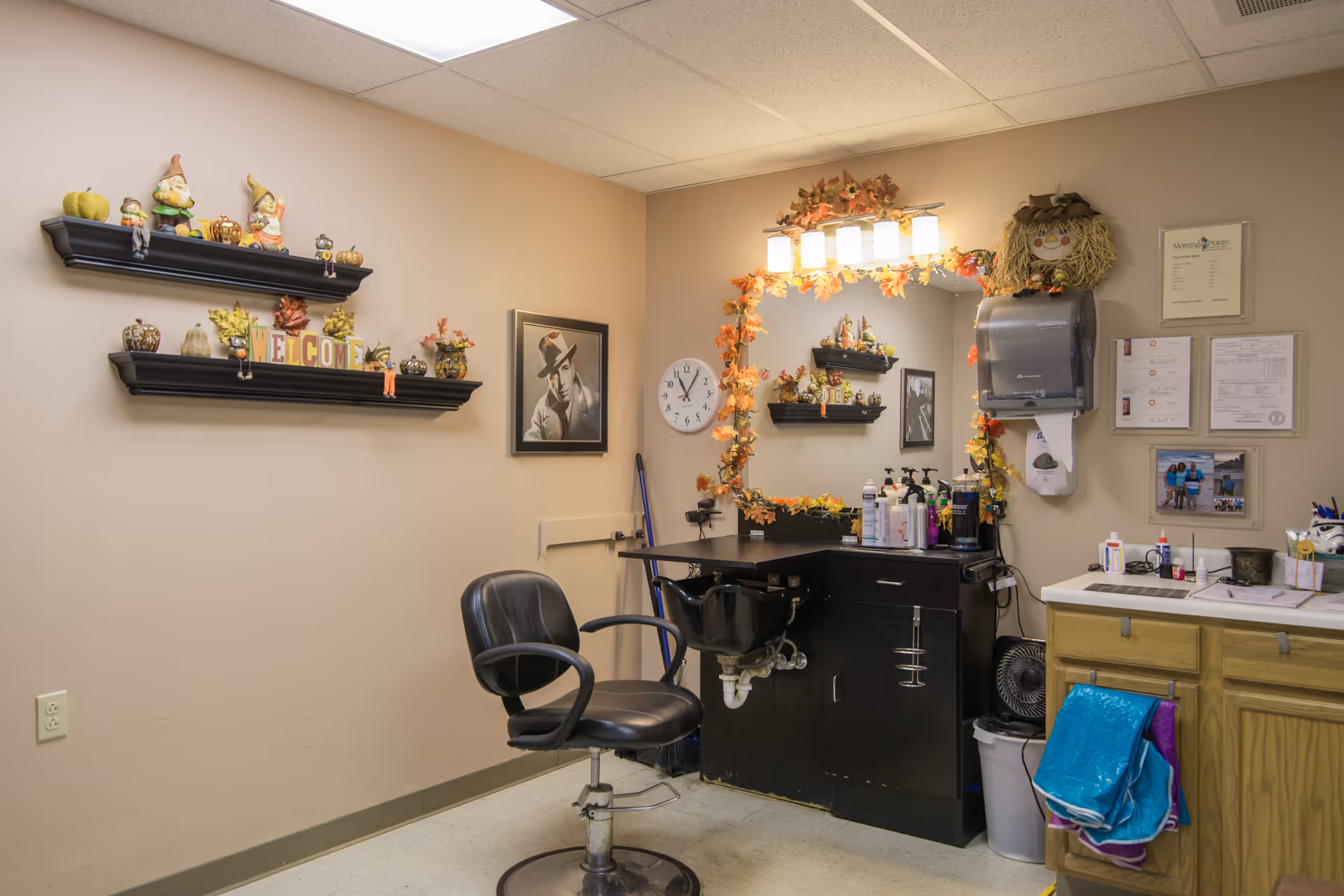 A small salon/styling station inside a senior facility with a barber chair, wash sink, mirror decorated with autumn garlands, and shelves of seasonal decor.
