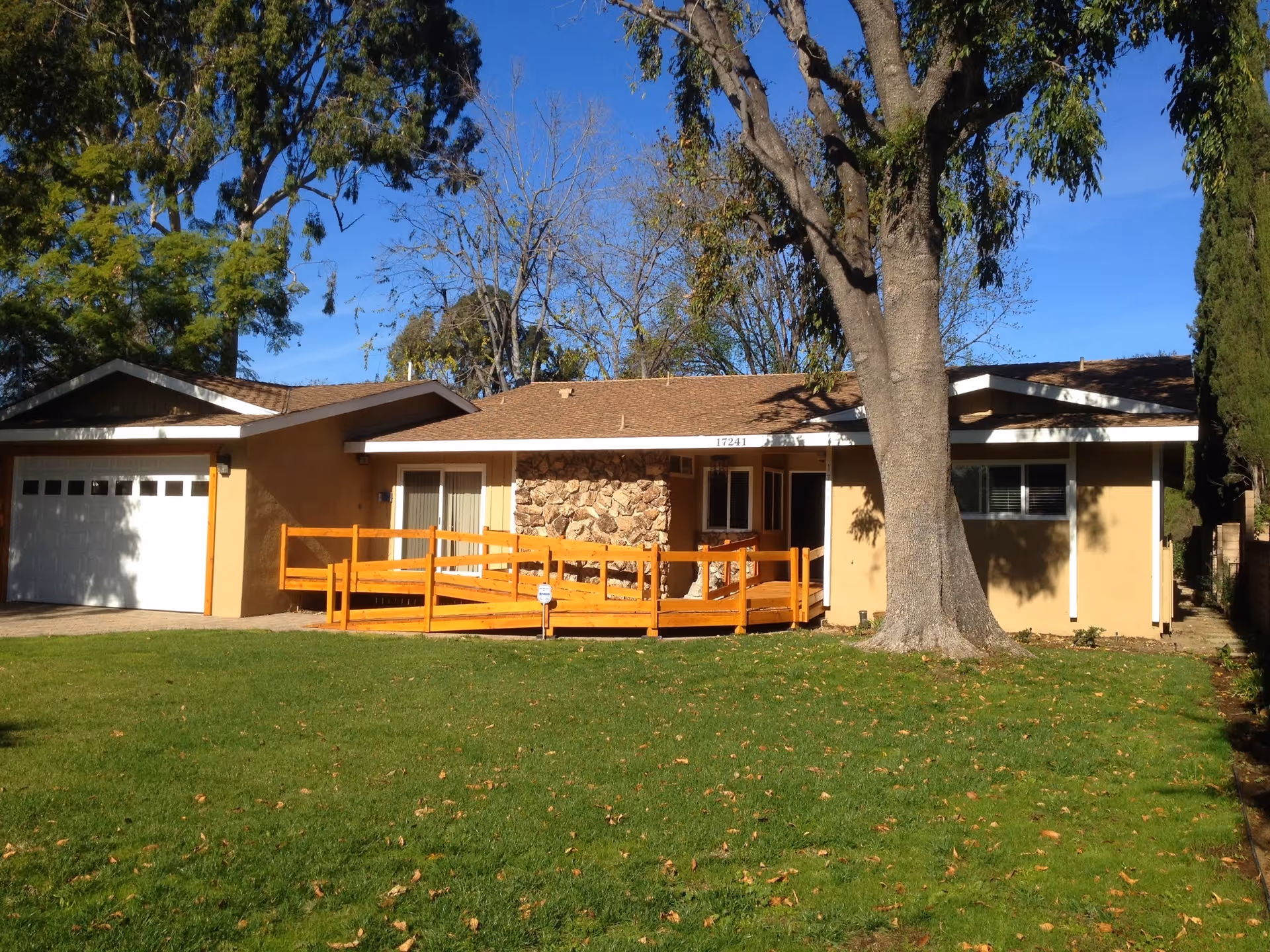 Single-story beige building with a wooden ramp, green lawn, and large tree.